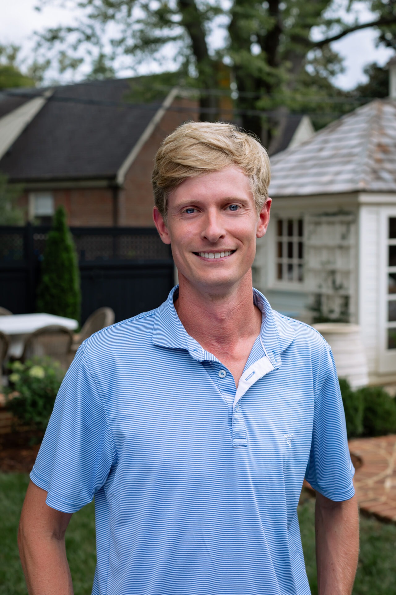 A person in a blue shirt smiles outside near a garden, with chairs, a table, and a small house visible in the background.