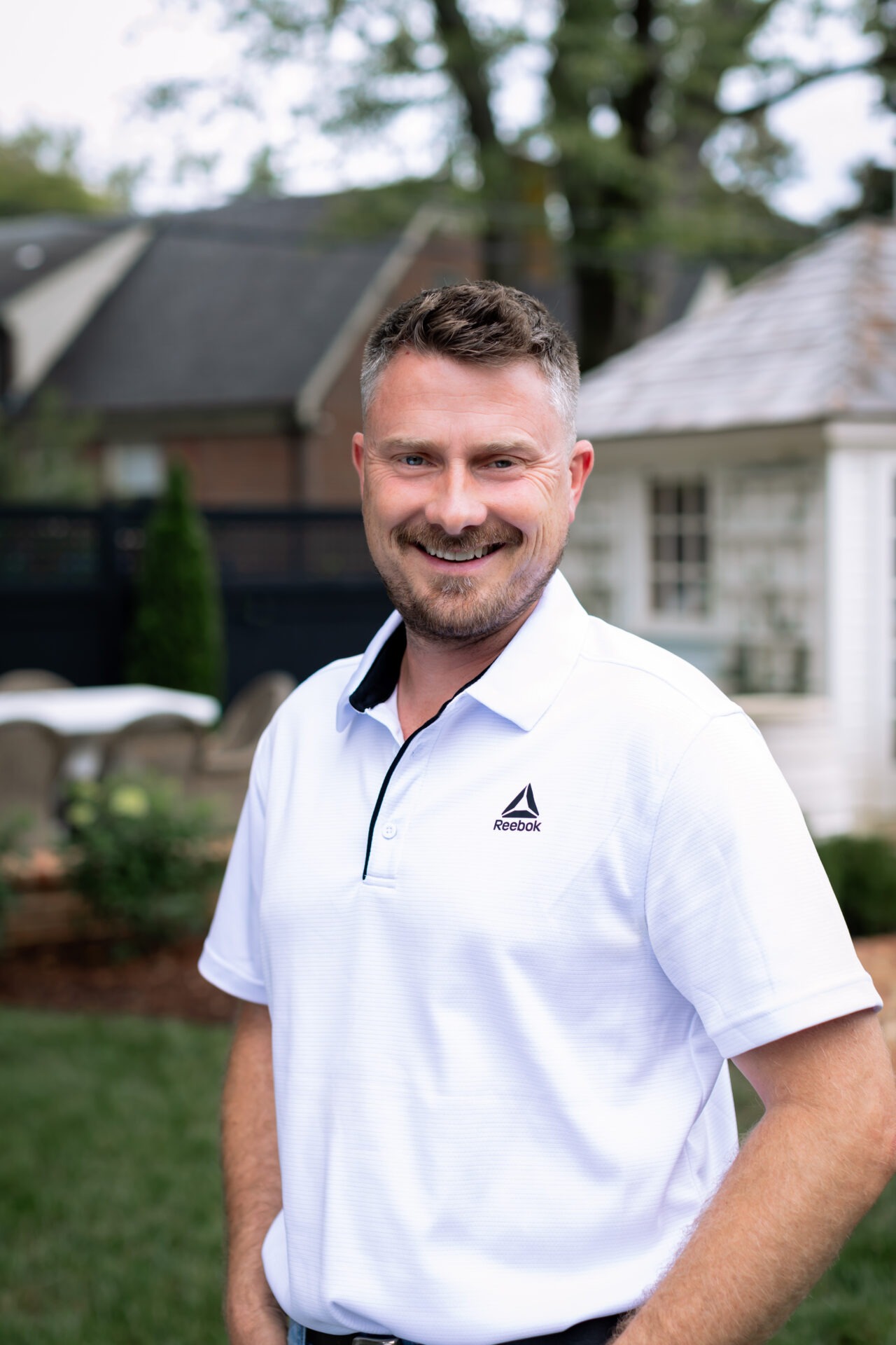 A smiling person in a white Reebok shirt stands outdoors. Background shows a small house and trees on a grassy yard.