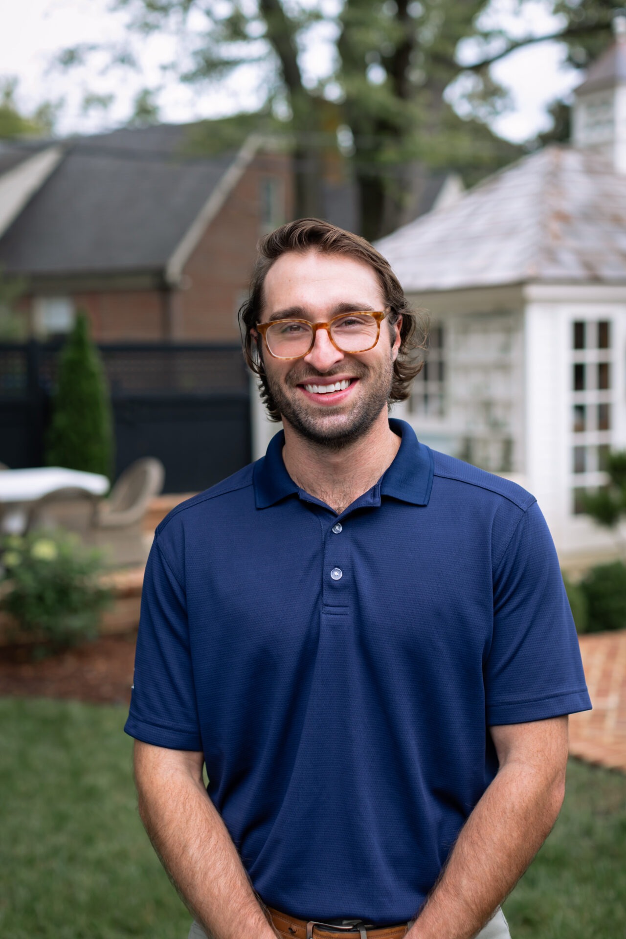 A person in glasses and a blue polo stands outdoors, smiling. Trees and a small building are visible in the background.