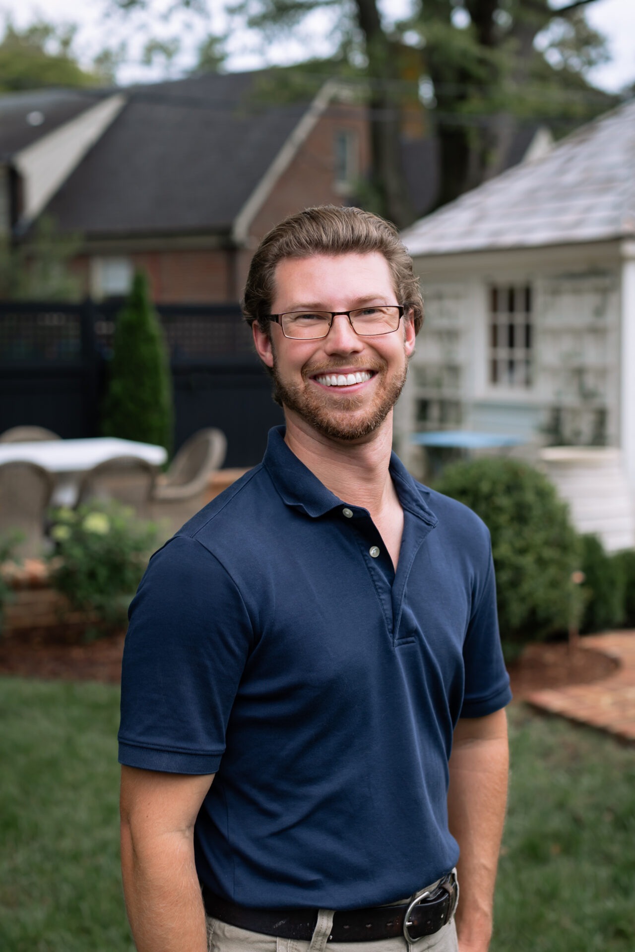 A person with glasses smiles outdoors, standing in a garden with a small white building and a seating area in the background.