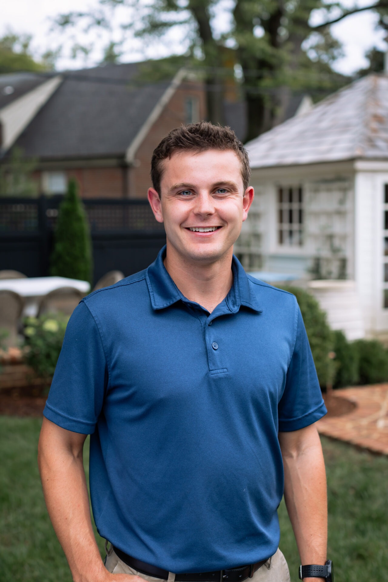 A person in a blue shirt stands smiling in a backyard, with trees, a house, and patio furniture in the background.