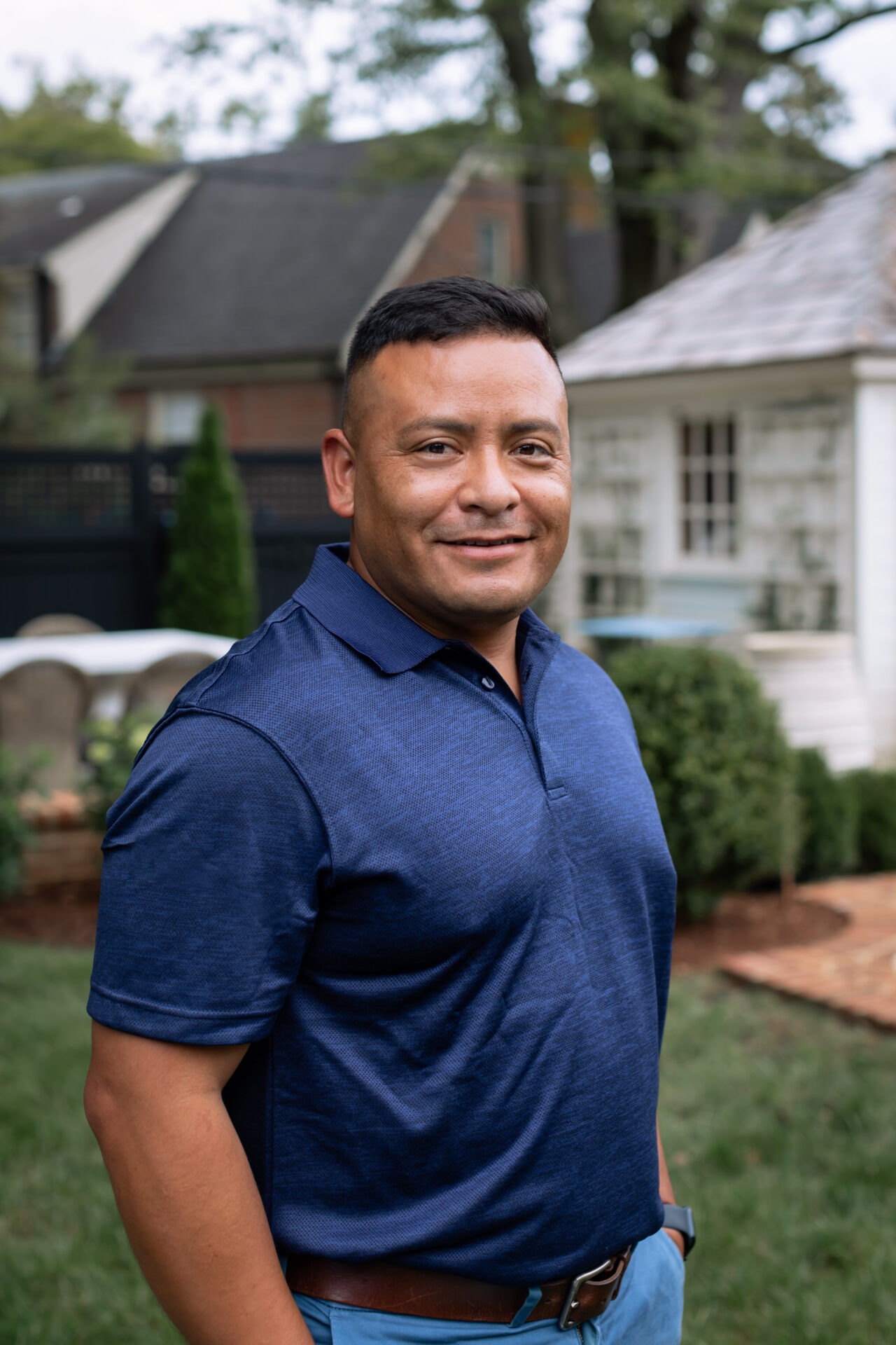 A person in a blue shirt stands in a garden setting with small buildings and greenery in the background.