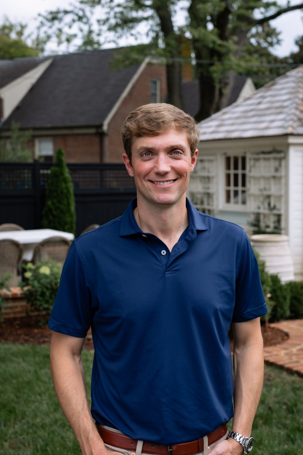 A person in a blue shirt stands smiling in a garden with a white shed, brick pathway, and trees in the background.