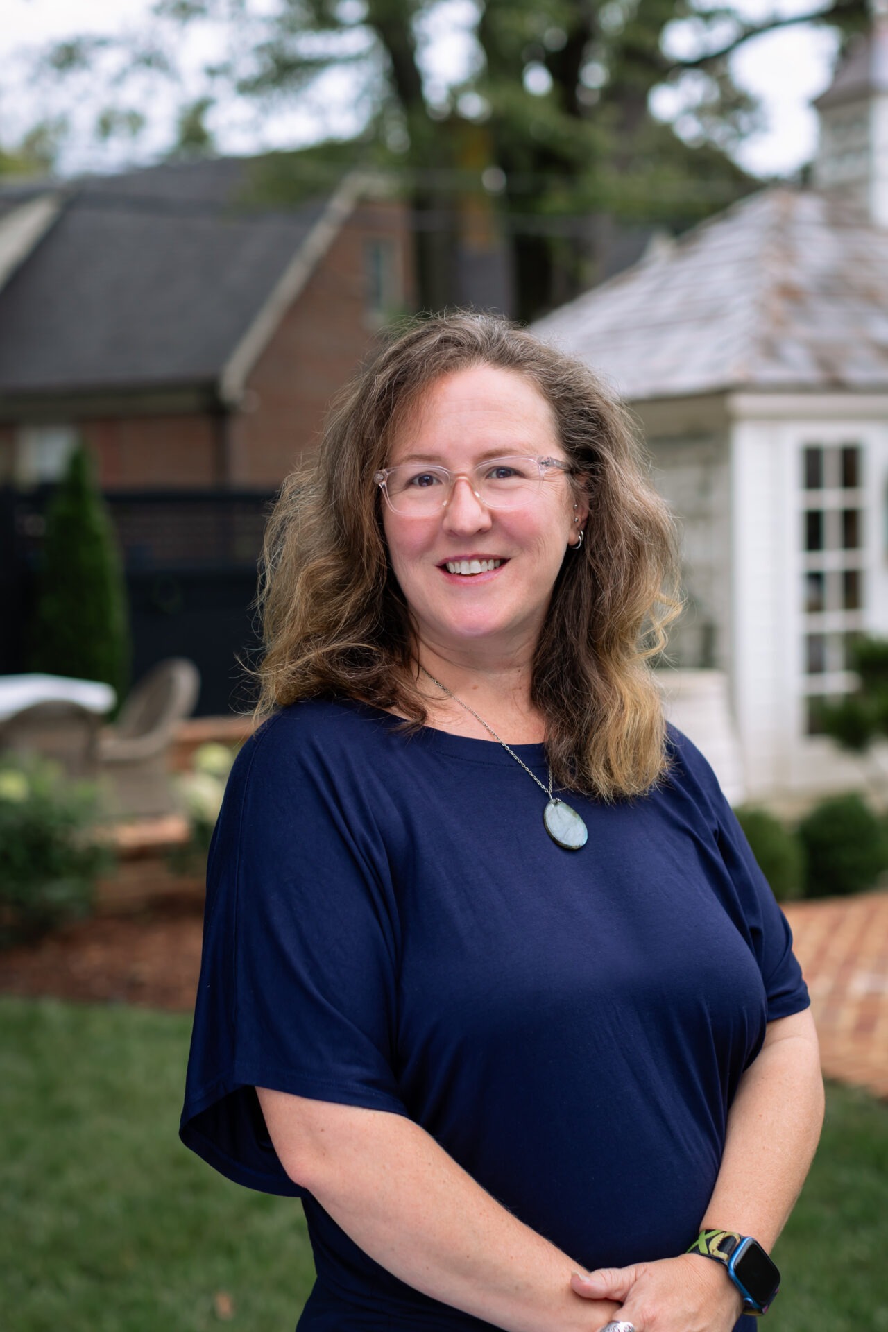 A person with long hair and glasses smiles while standing in a garden with stone buildings in the background.