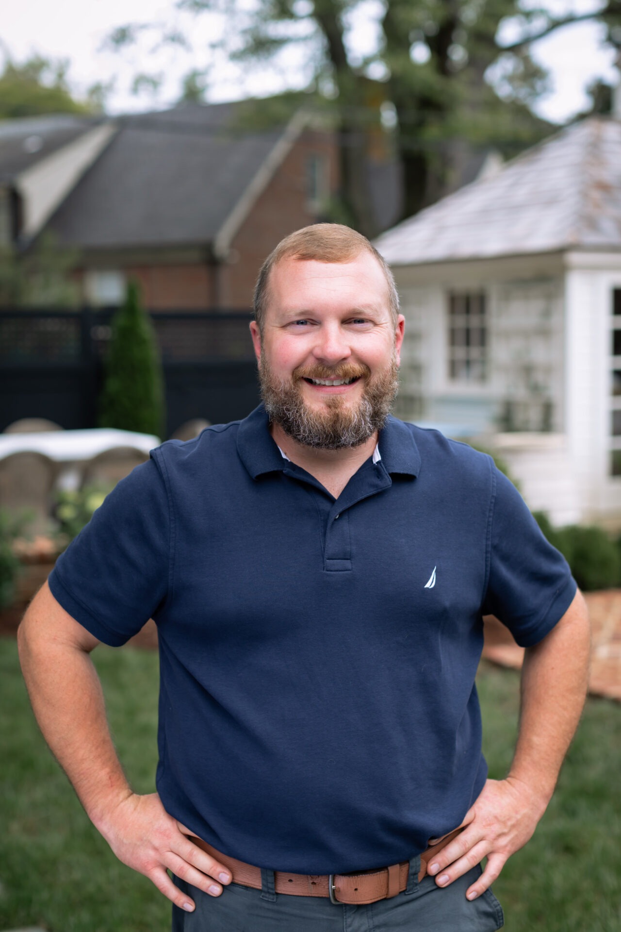 A person in a blue shirt stands smiling in a garden with houses and trees in the background.