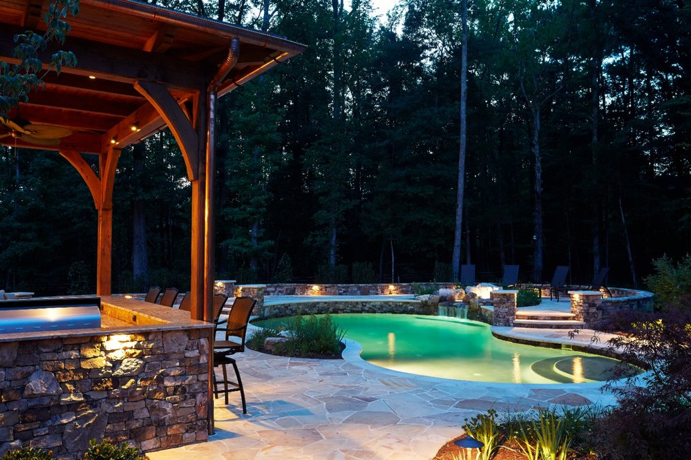 A serene backyard setting features a glowing pool, stone patio, wooden pergola, and surrounding trees at dusk, creating a tranquil evening ambiance.