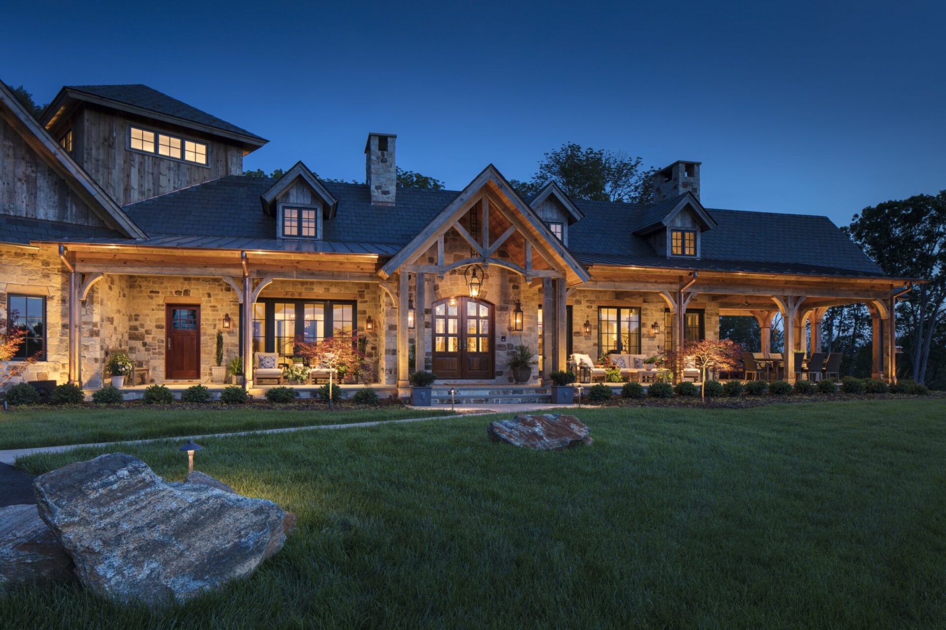 A cozy, rustic house with stone and wood exteriors is illuminated at dusk, surrounded by a manicured lawn and trees in the background.