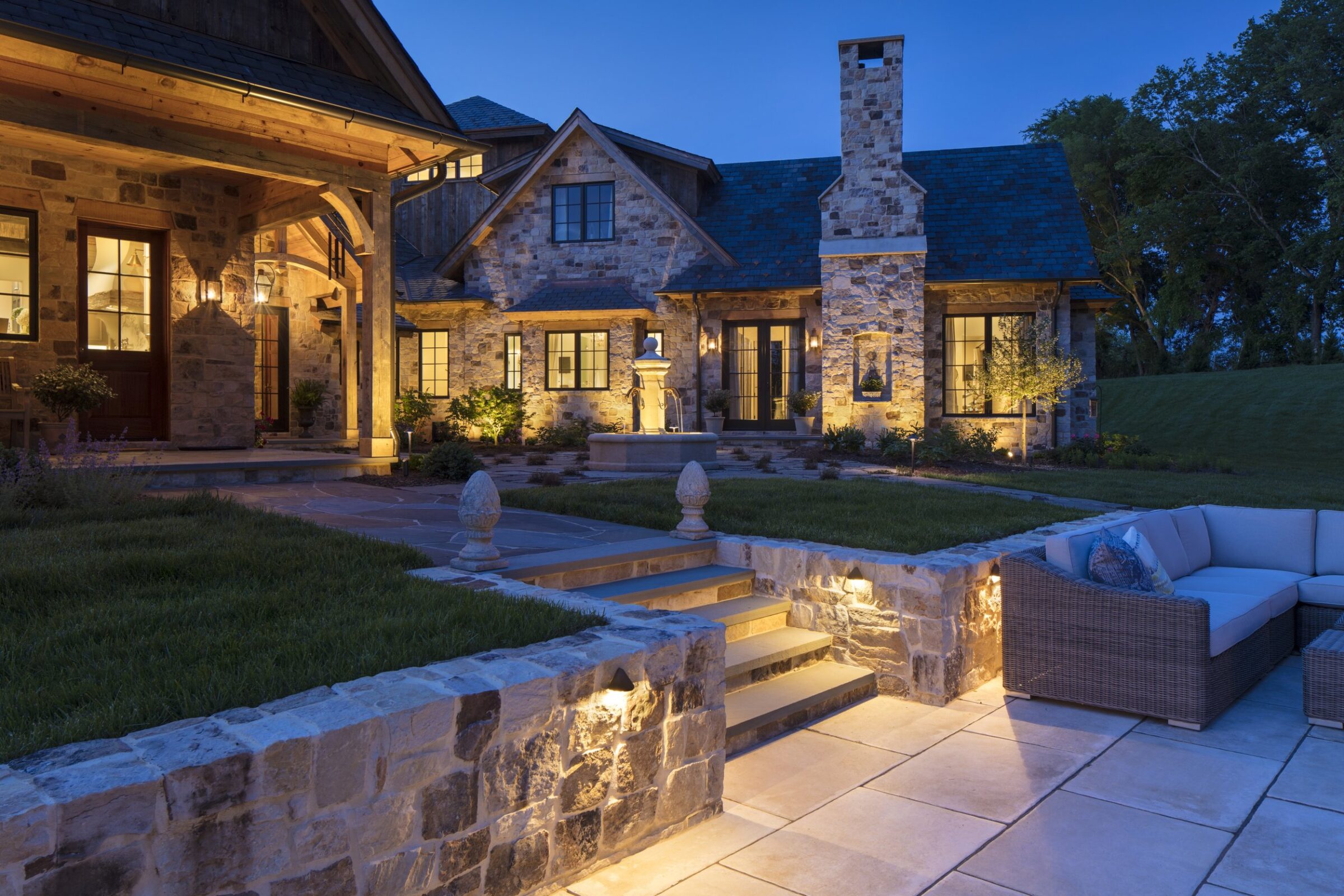 A stone house with warm exterior lighting at dusk, featuring a cozy outdoor seating area and beautifully landscaped garden steps.