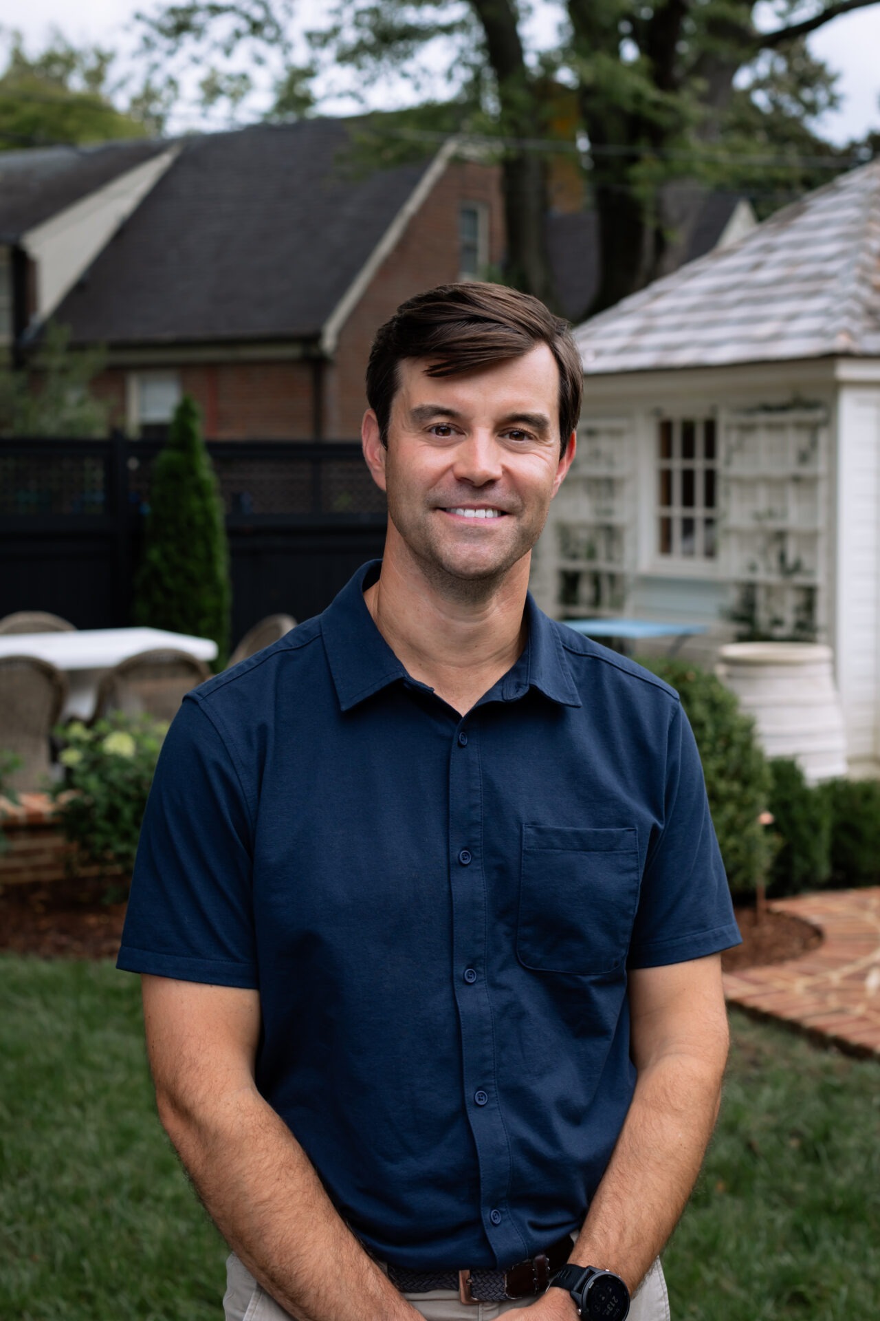 A person in a blue shirt stands in a garden with a small house and trees in the background.