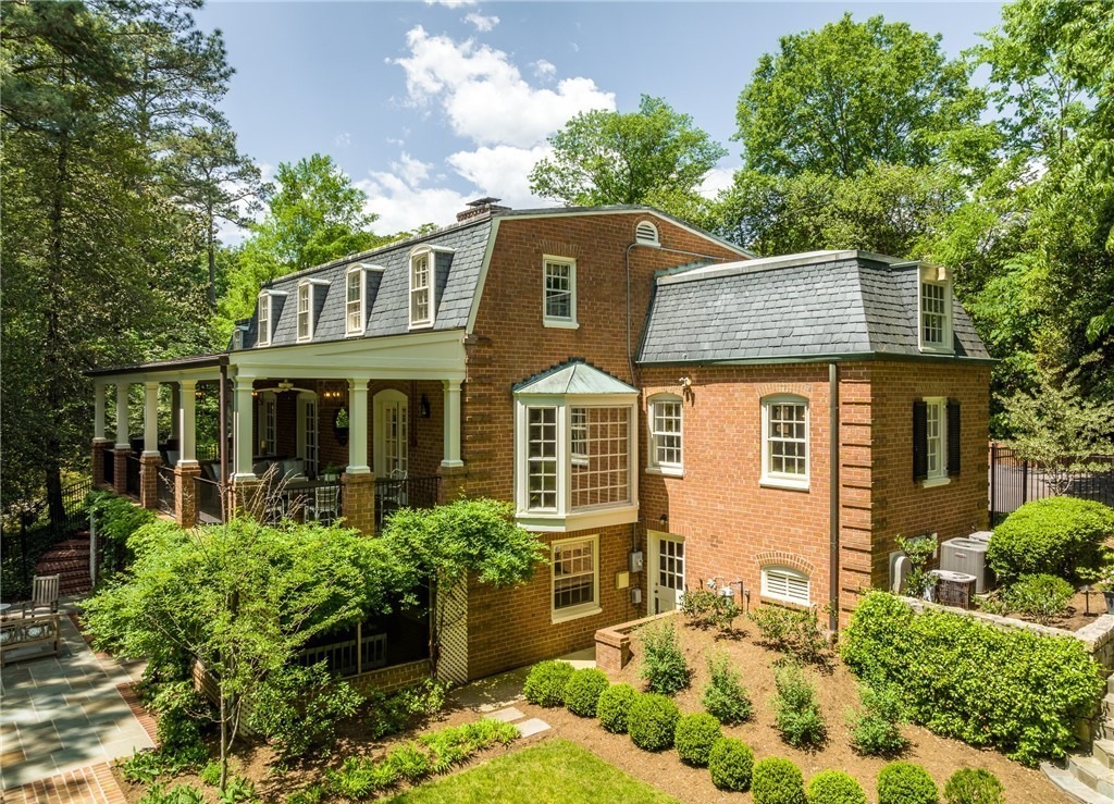 A large brick colonial-style house with a wraparound porch, surrounded by lush greenery and trees, under a clear, partly cloudy sky.