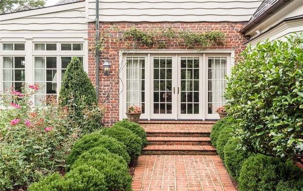 Brick house with white-framed glass doors, surrounded by greenery and blooming flowers. Steps lead to entrance; cozy, inviting garden setting.