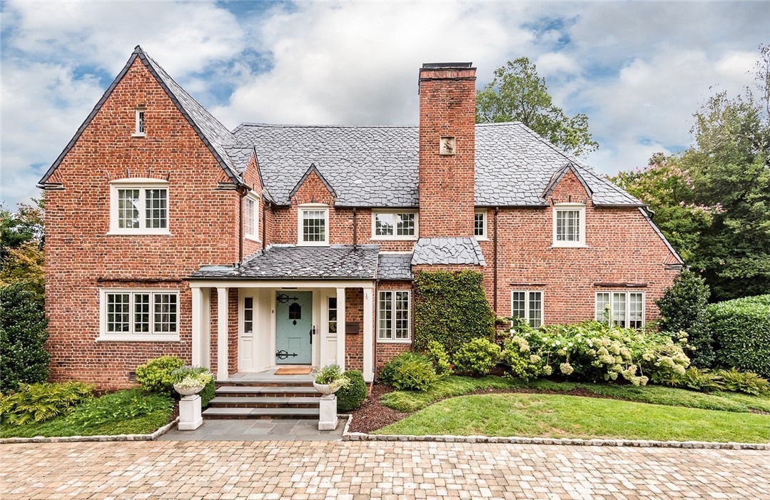 A charming two-story brick house with slate roof, lush greenery, and manicured grounds. Features white-framed windows and a prominent central chimney.