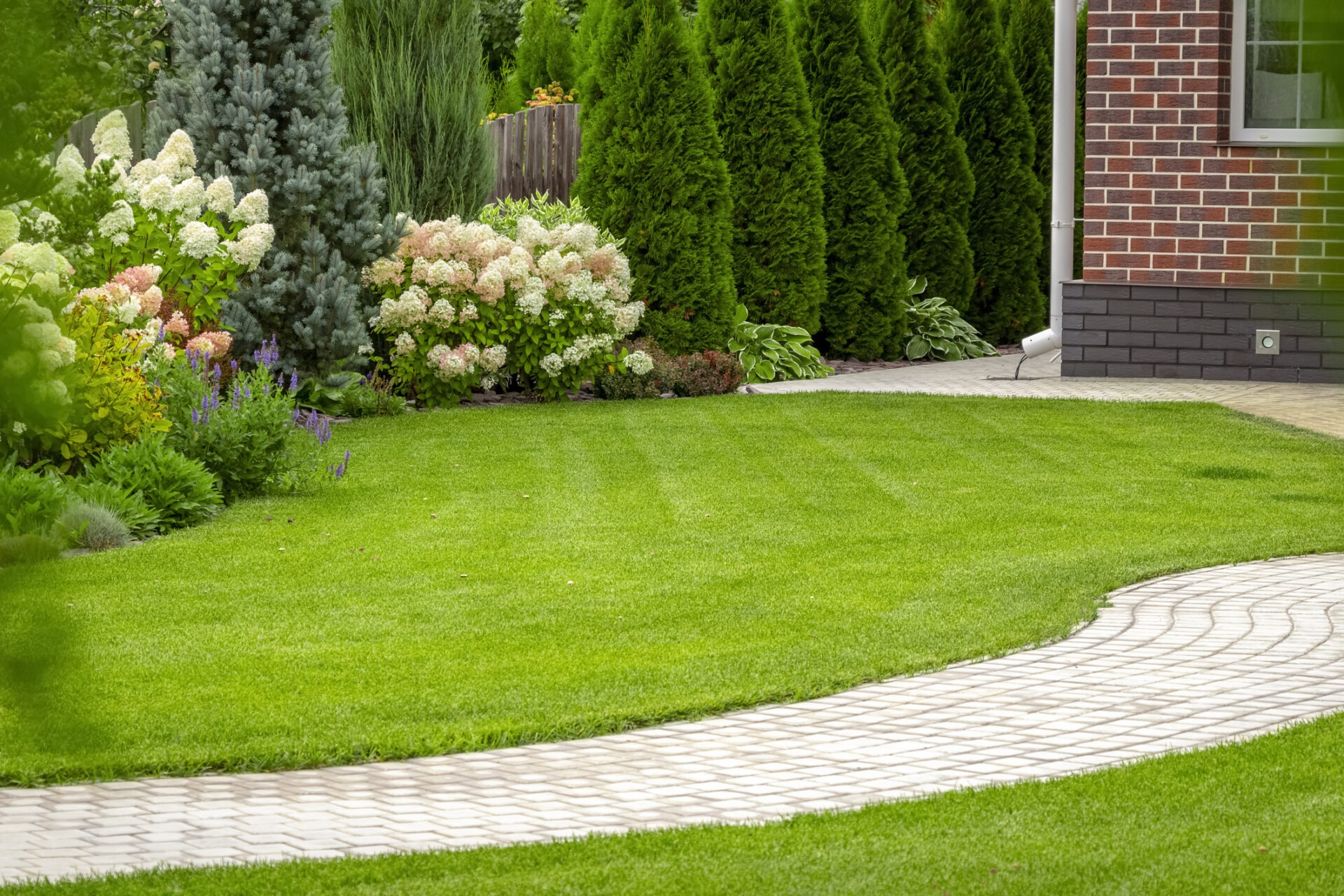 A lush garden with neatly trimmed grass and blooming flowers alongside a brick building and a curving stone pathway.