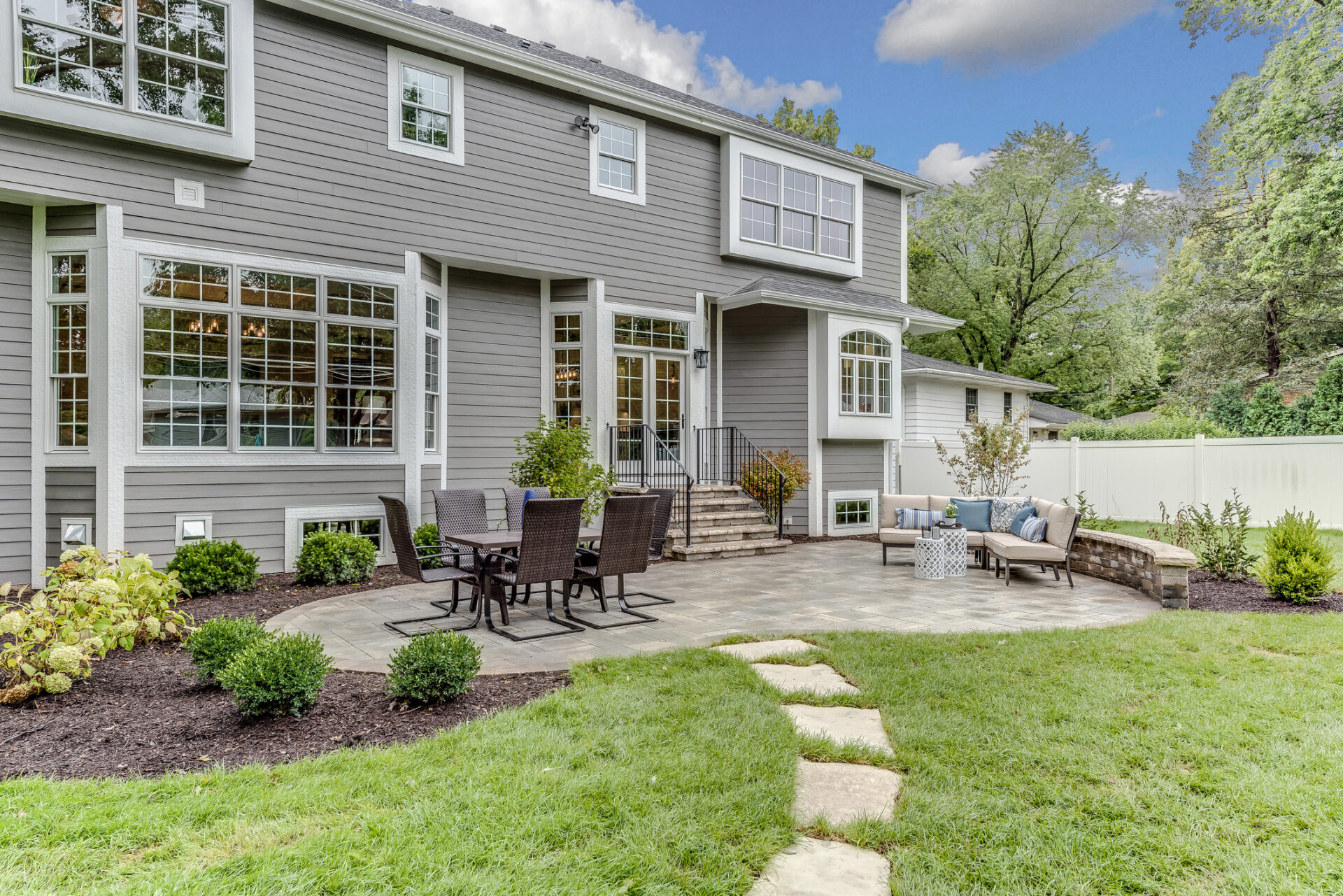 Spacious suburban house with gray siding, featuring a large outdoor patio with chairs and a table, surrounded by lush greenery and trees.