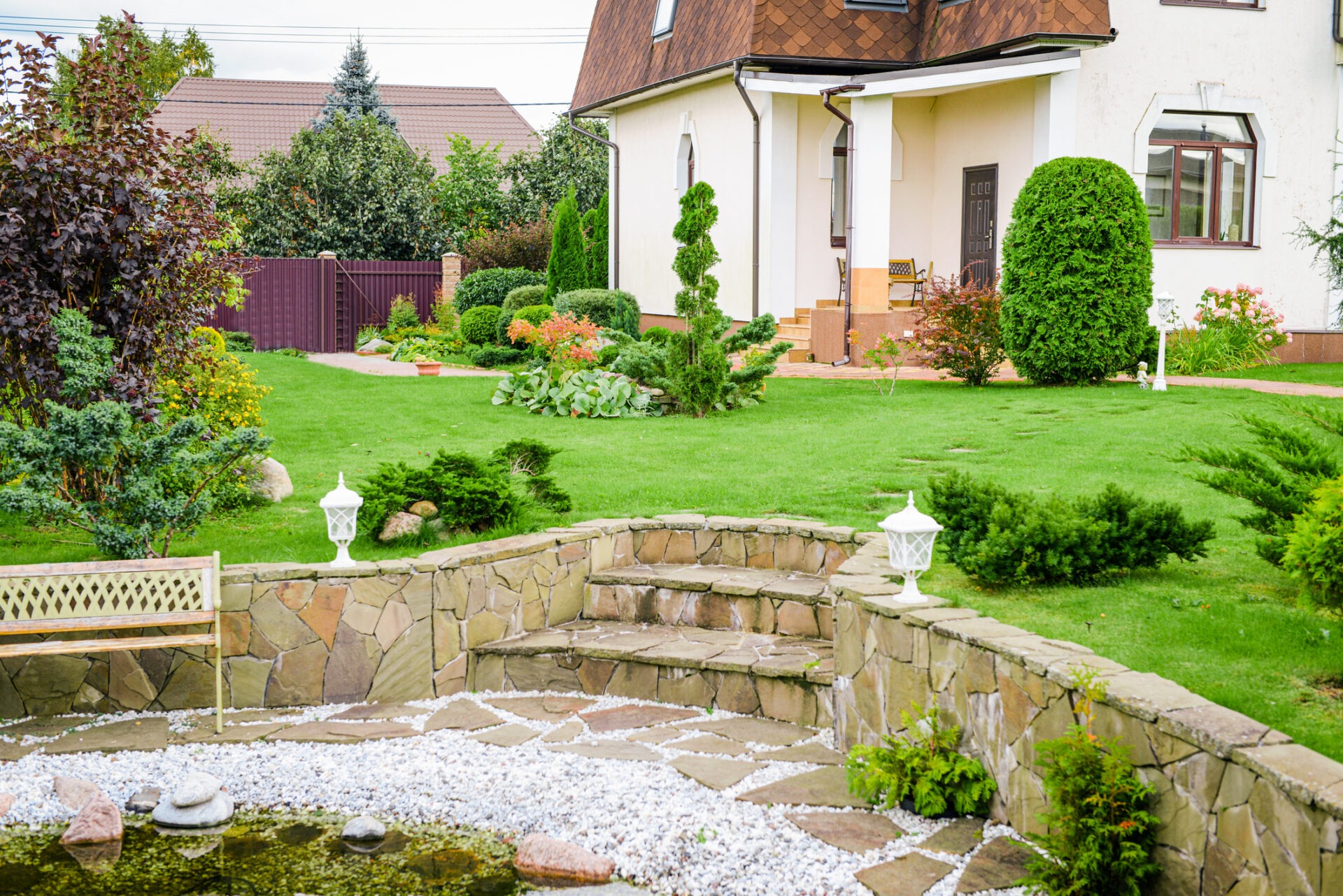 Charming garden with well-maintained greenery and stone pathway, adjoining a quaint house. Decorative lanterns and small pond enhance the peaceful ambiance.