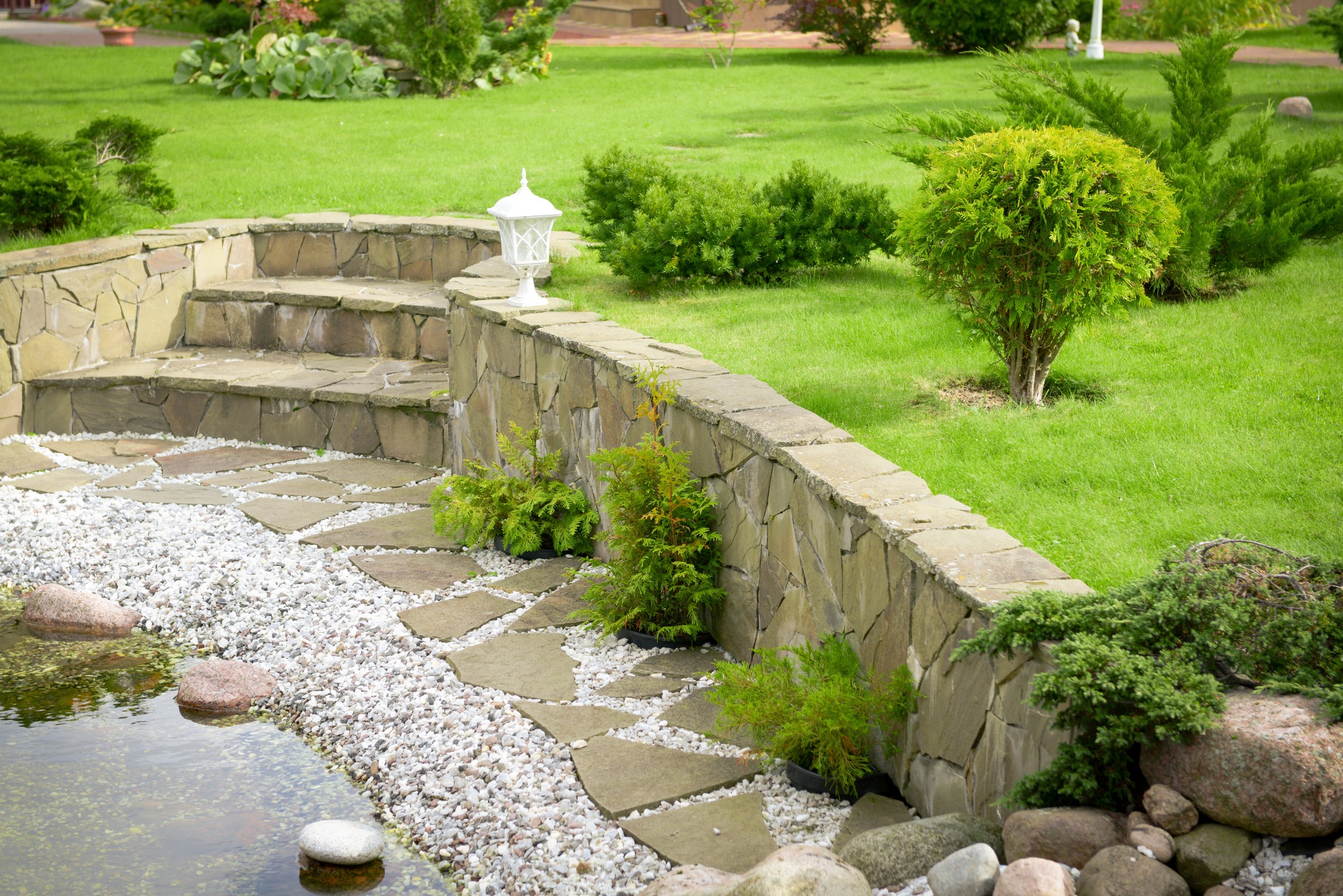 A serene garden with stone steps, a small pond, greenery, and a white lantern, surrounded by neatly trimmed bushes and an inviting landscape.