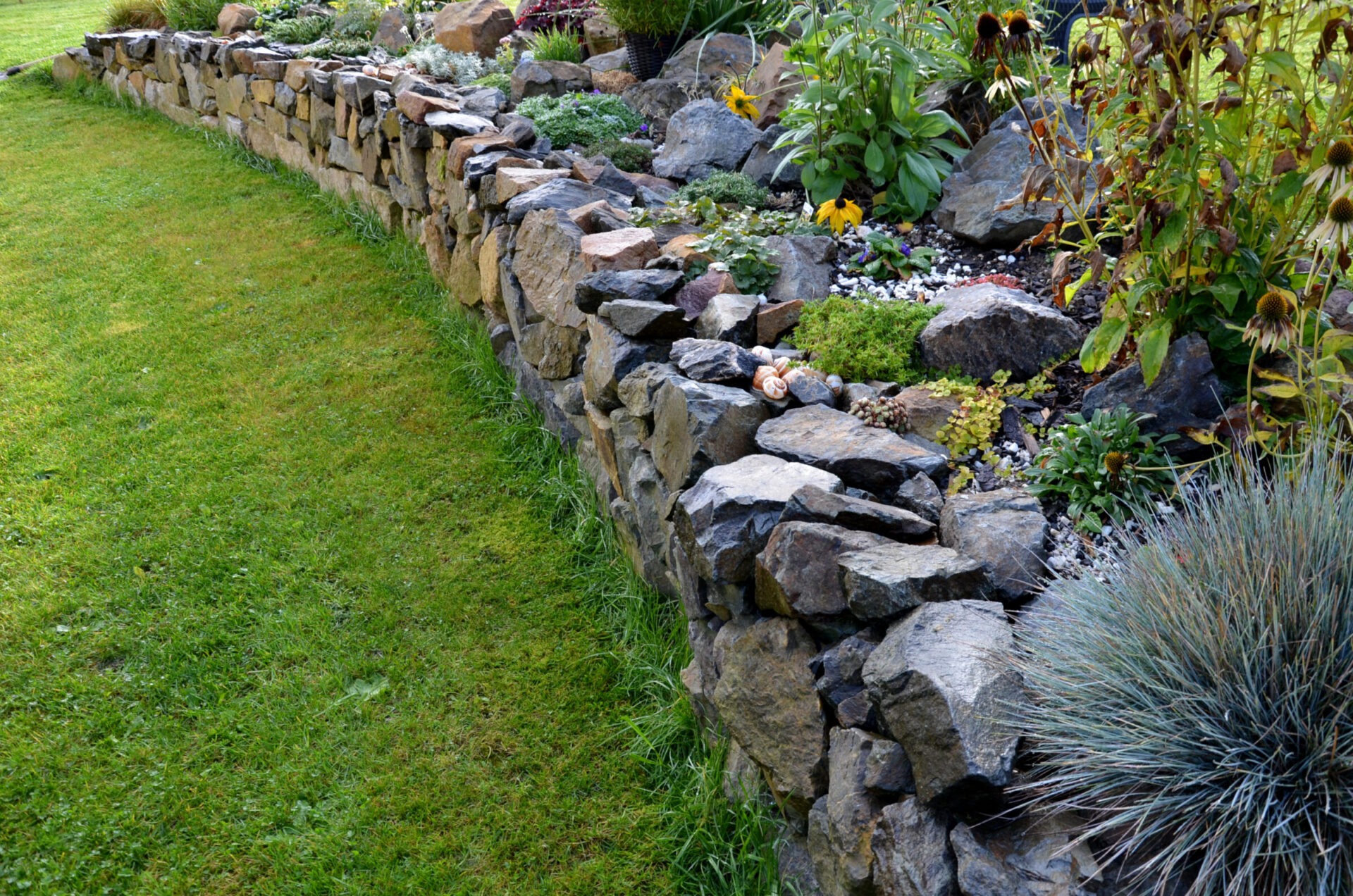 Stone wall borders a lush garden with various plants and flowers. The grass is neatly trimmed, and there are no people or buildings visible.