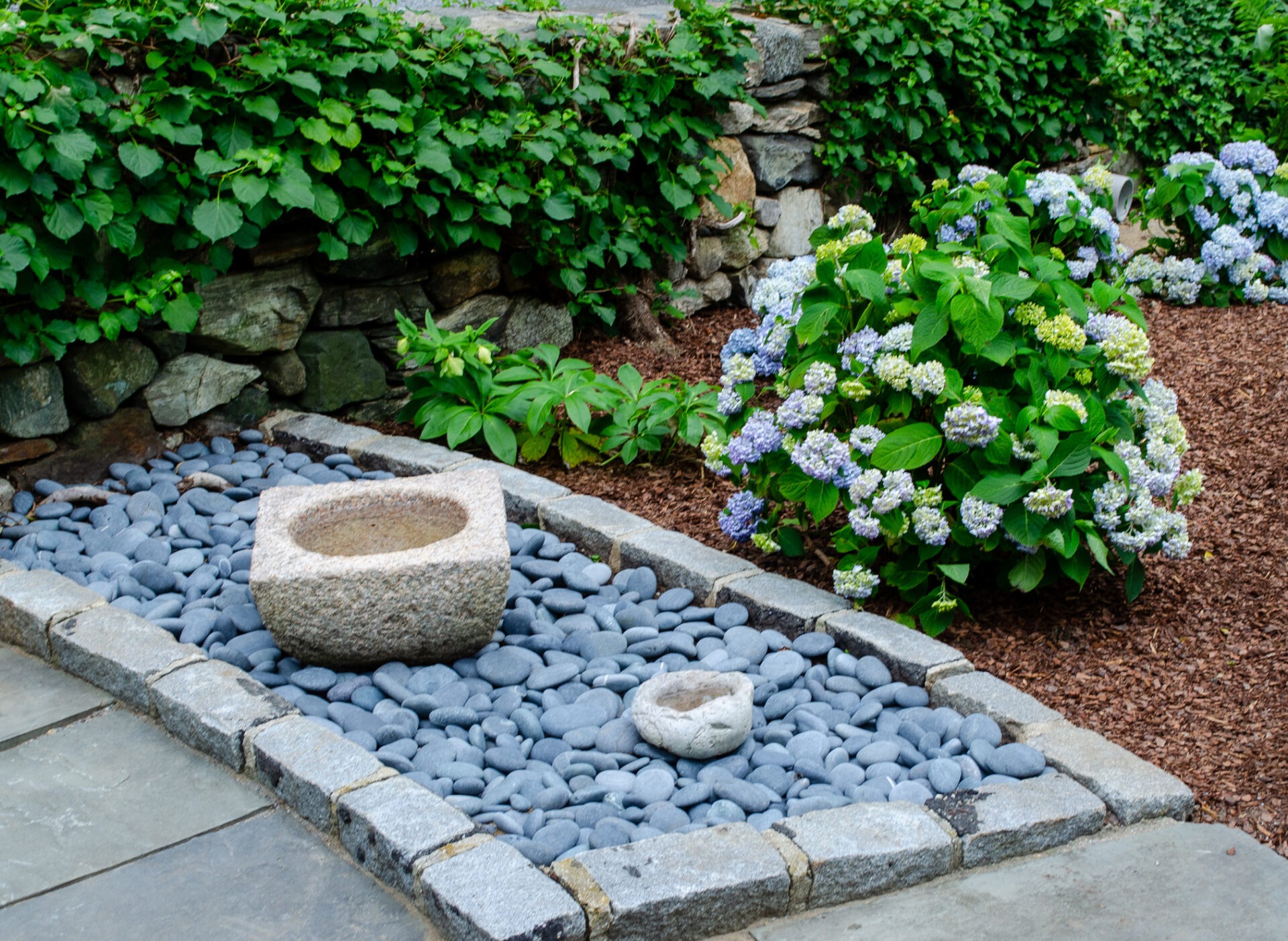 A serene garden with stone pathways, a stone bowl, smooth rocks, and blooming hydrangeas, surrounded by a lush green hedge.