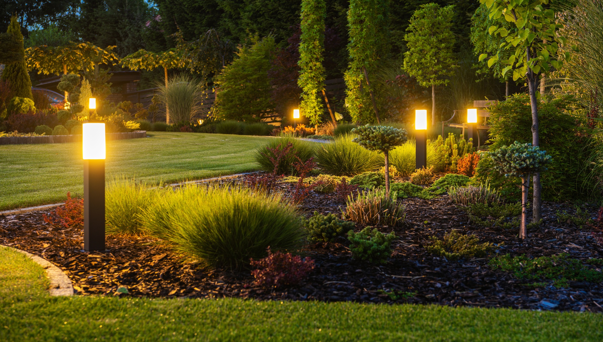 A beautifully landscaped garden at dusk, featuring illuminated pathway lights, neatly trimmed bushes, lush greenery, and a serene, peaceful ambiance.