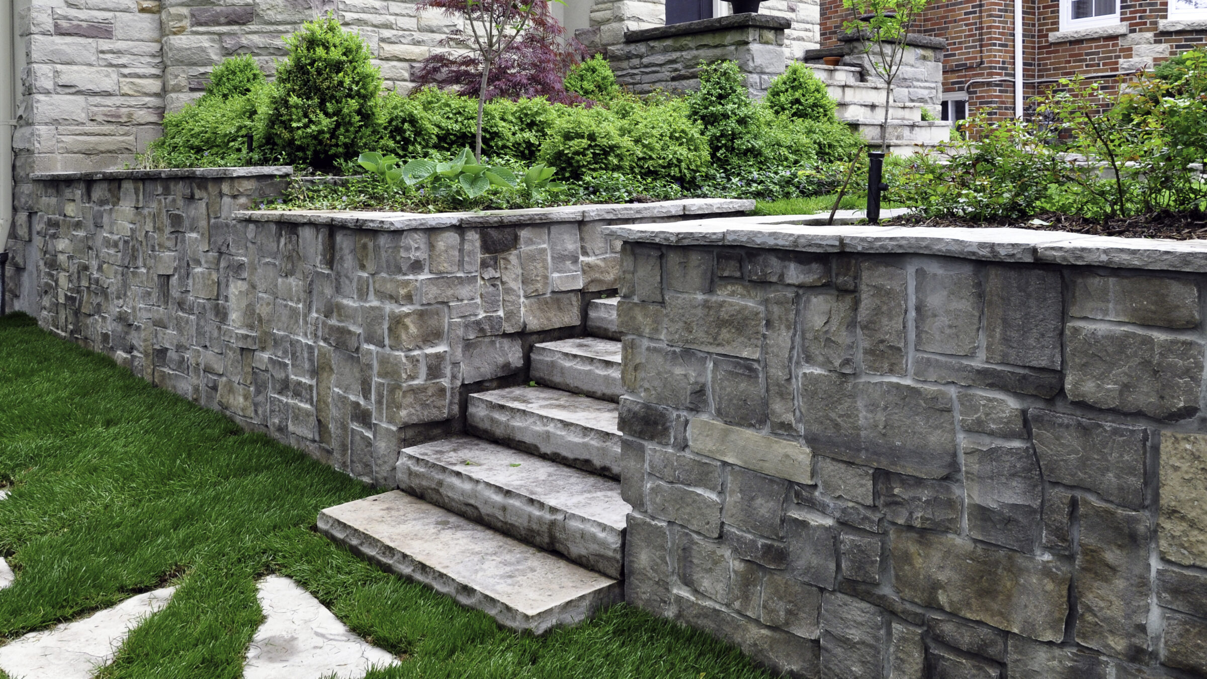 Stone steps and walls with greenery lead to a residence. Trimmed grass surrounds the area, creating a neat and inviting outdoor space.