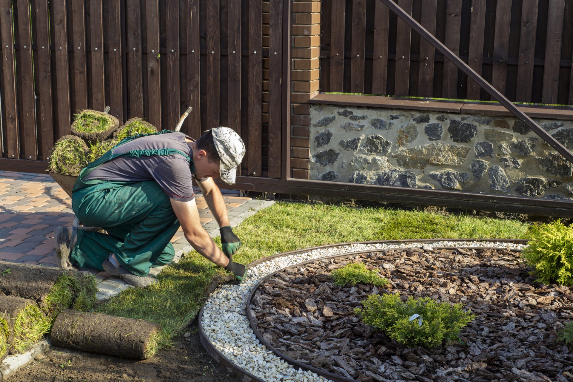 A person in work attire is laying sod in a landscaped garden area surrounded by a wooden fence and stone wall.