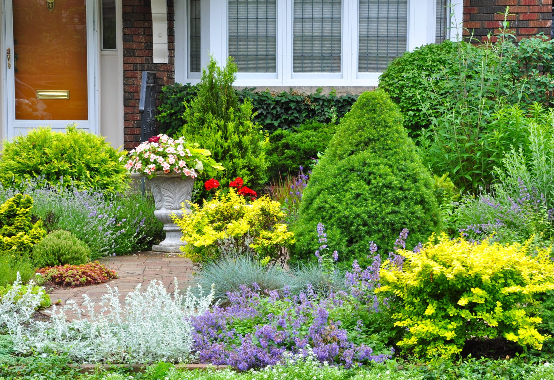 A lush garden with diverse colorful flowers and shrubs decorates the entrance of a brick house with white-framed windows and a wooden door.