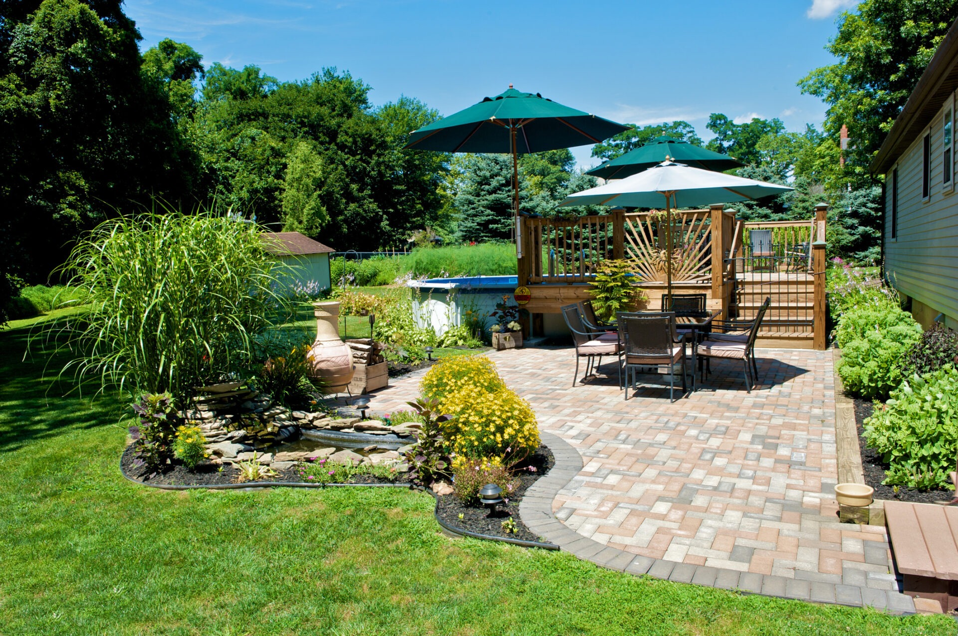 A landscaped backyard features a patio with tables, umbrellas, and a small pond, surrounded by lush greenery and a wooden fence.