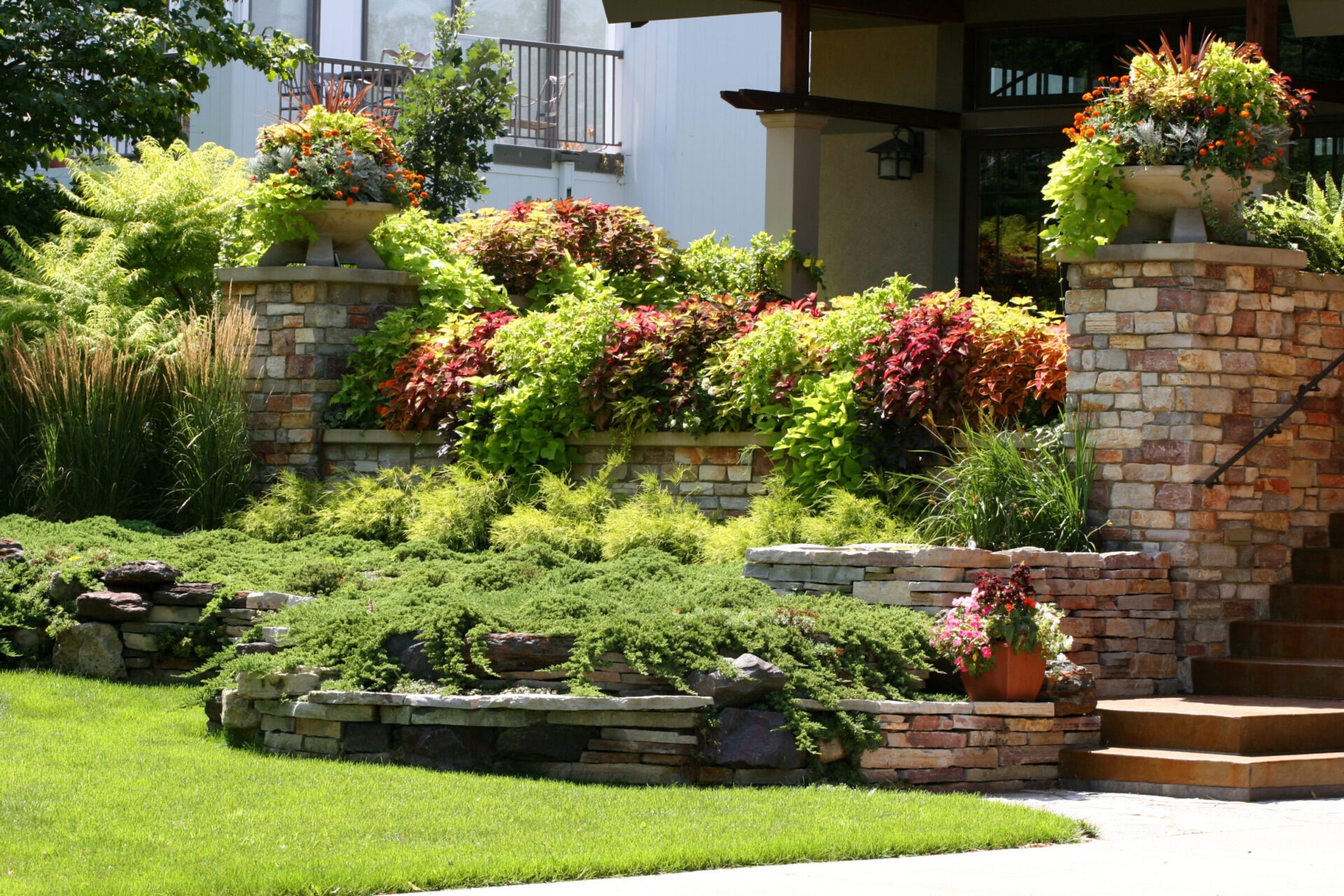 A lush, landscaped garden with stone walls, vibrant plants, and steps leading to a building entrance; no people or landmarks visible.