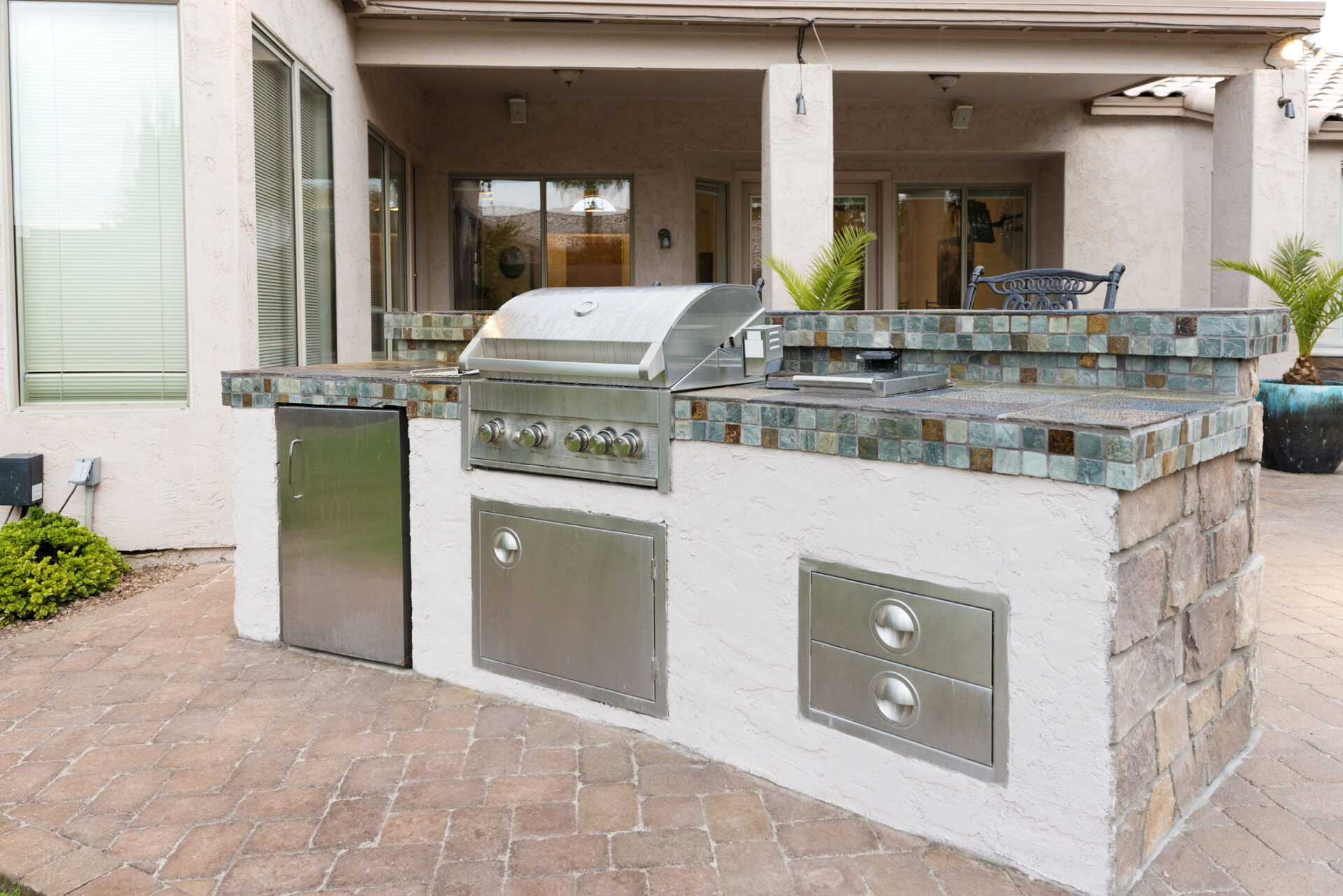 Outdoor kitchen featuring a stainless steel grill and storage drawers, set on a stone patio near a modern residential building.