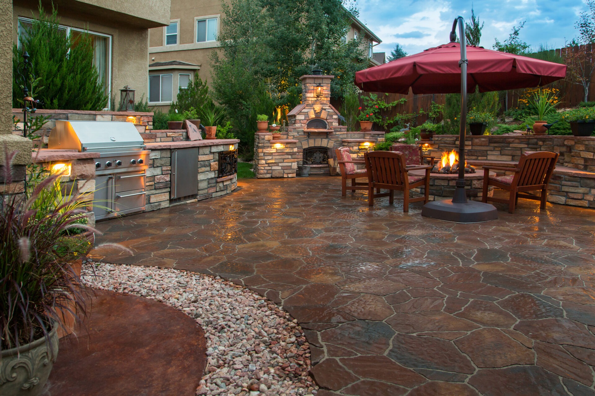 A cozy backyard with a stone patio, outdoor kitchen, fireplace, chairs, and umbrella, surrounded by plants and two house walls.