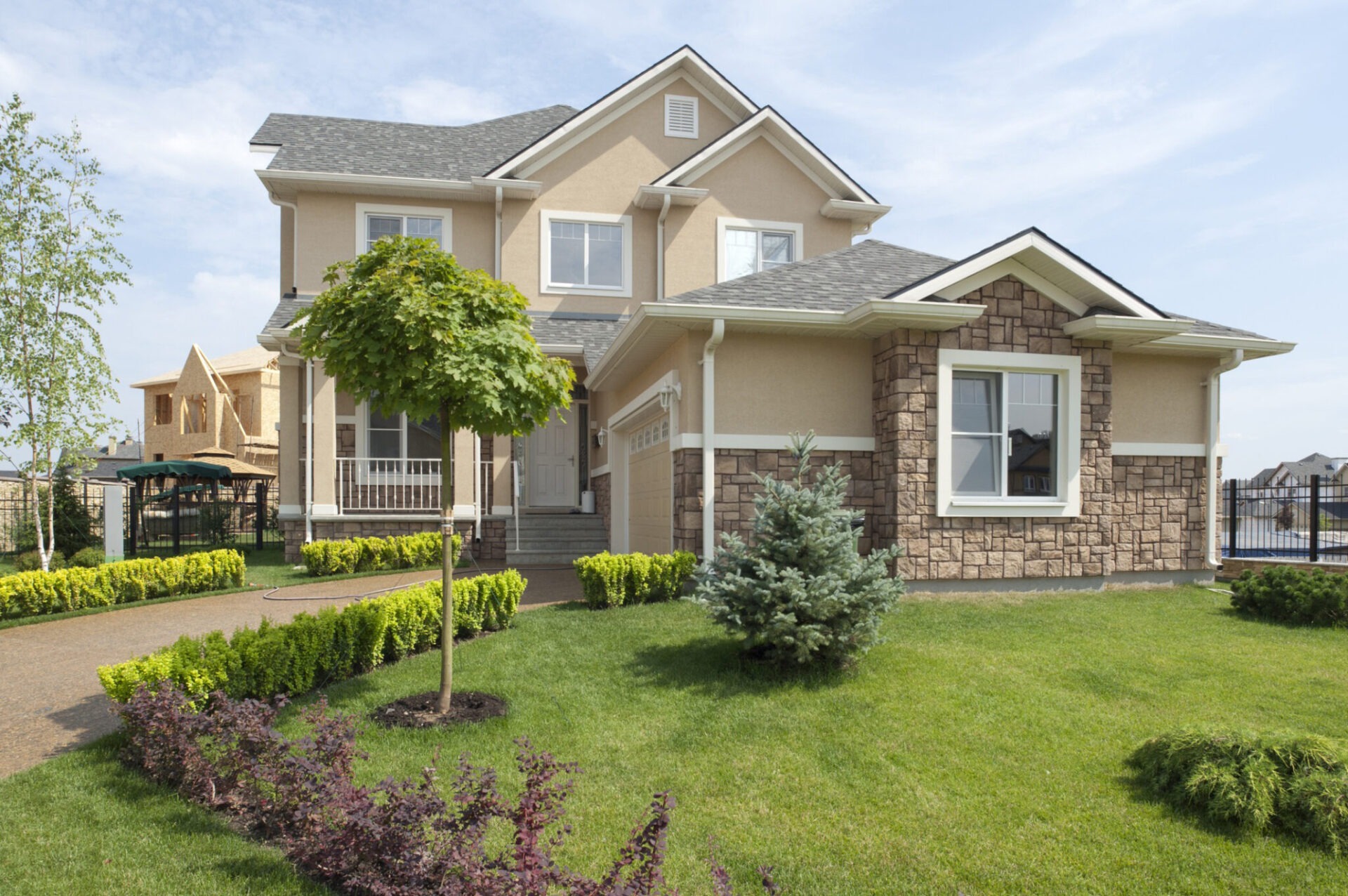 A two-story house with stone accents, neatly trimmed lawn, and a tree in the yard under a clear sky. No recognizable landmarks visible.