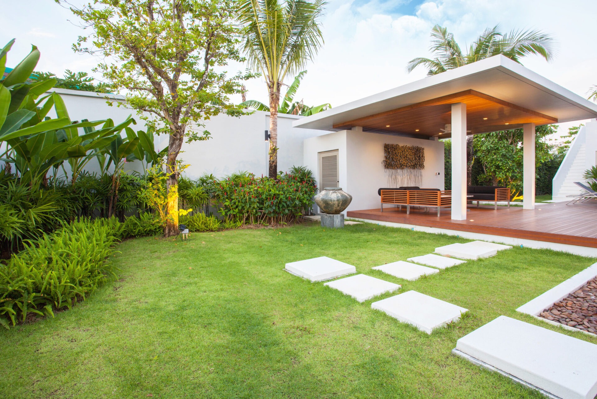 Modern garden patio with wooden deck, white pillars, lush plants, and stepping stones on manicured lawn, under clear skies and palm trees.
