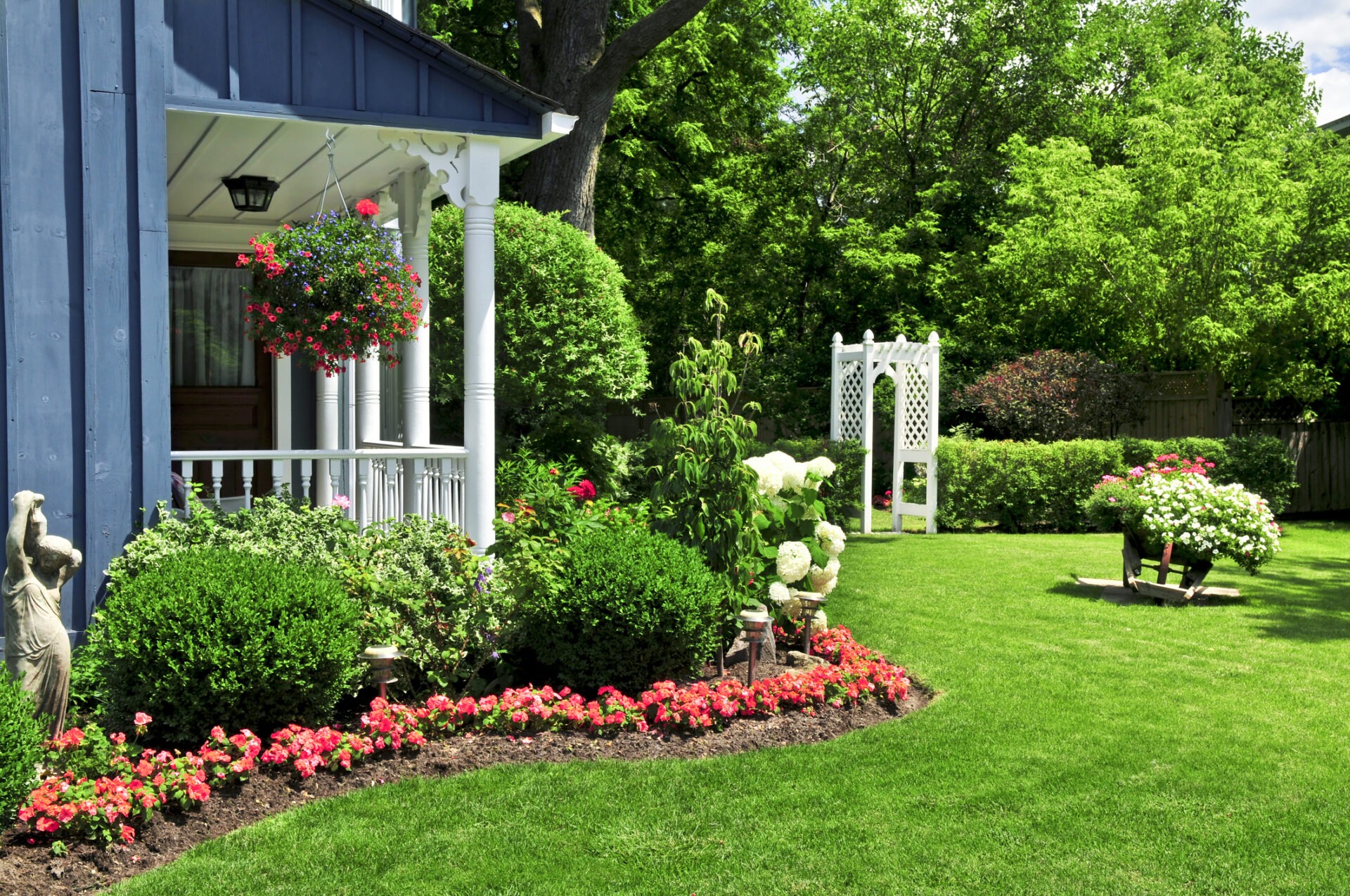 A serene garden with lush greenery, colorful flowers, a white pergola, and a charming blue house with a front porch adorned with hanging baskets.