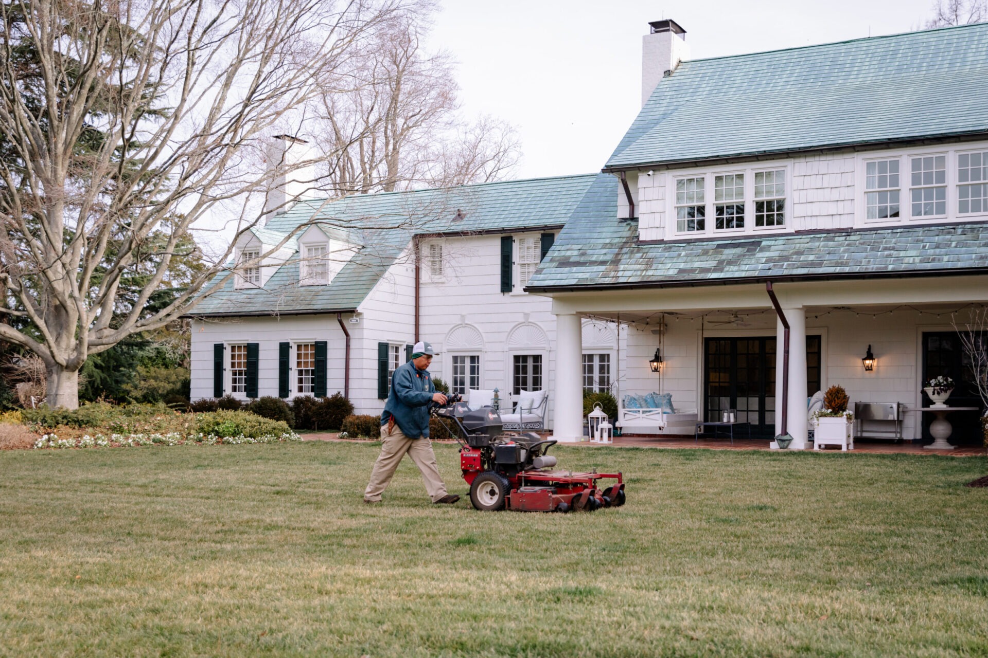 A person operates a lawn mower on a well-kept lawn in front of a large, white, two-story house with a green roof.