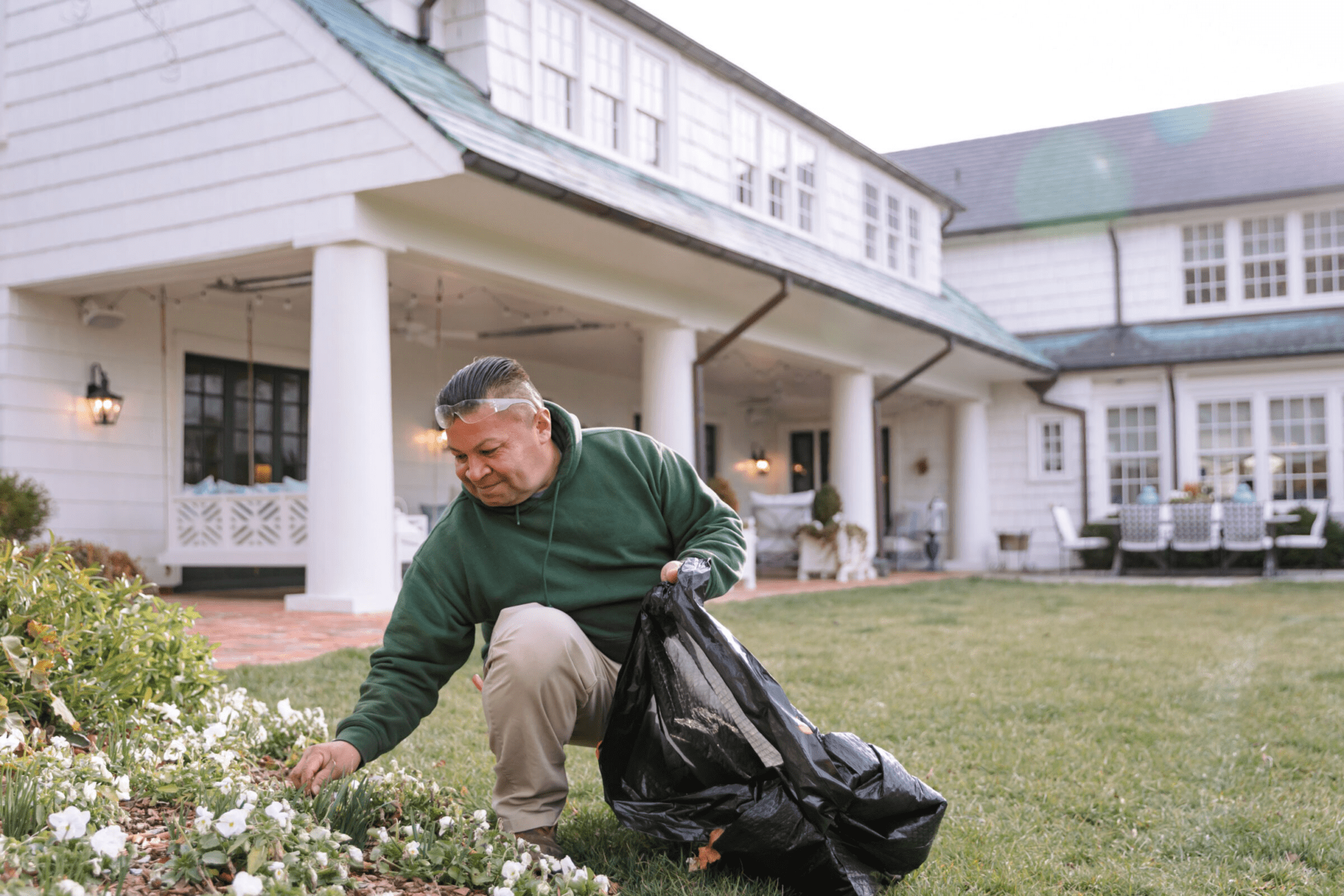 A person wearing a green hoodie tends to a garden in front of a large, white, colonial-style house with columns and multiple windows.