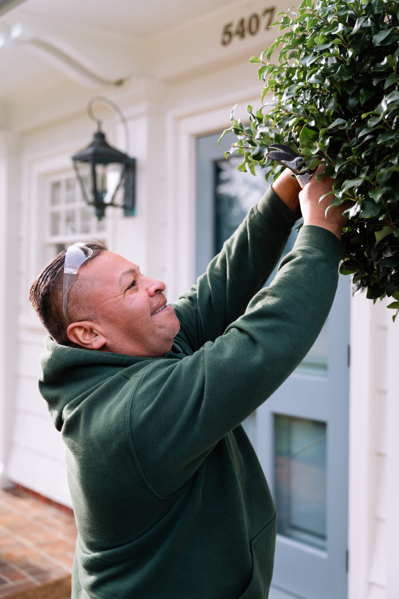 A person in a green hoodie trims a bush outside a house with a door and lantern. House number "5407" is visible above.