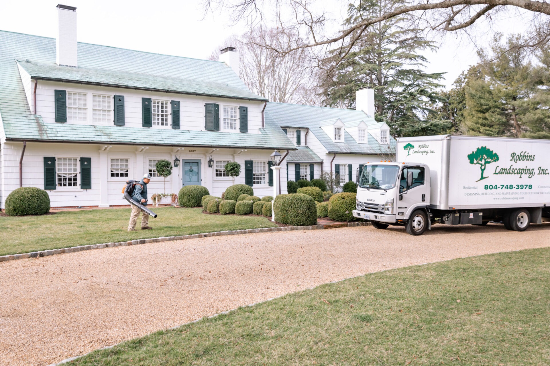 A person using landscaping equipment in front of a large house. A truck labeled “Robbins Landscaping, Inc.” is parked nearby on a gravel driveway.