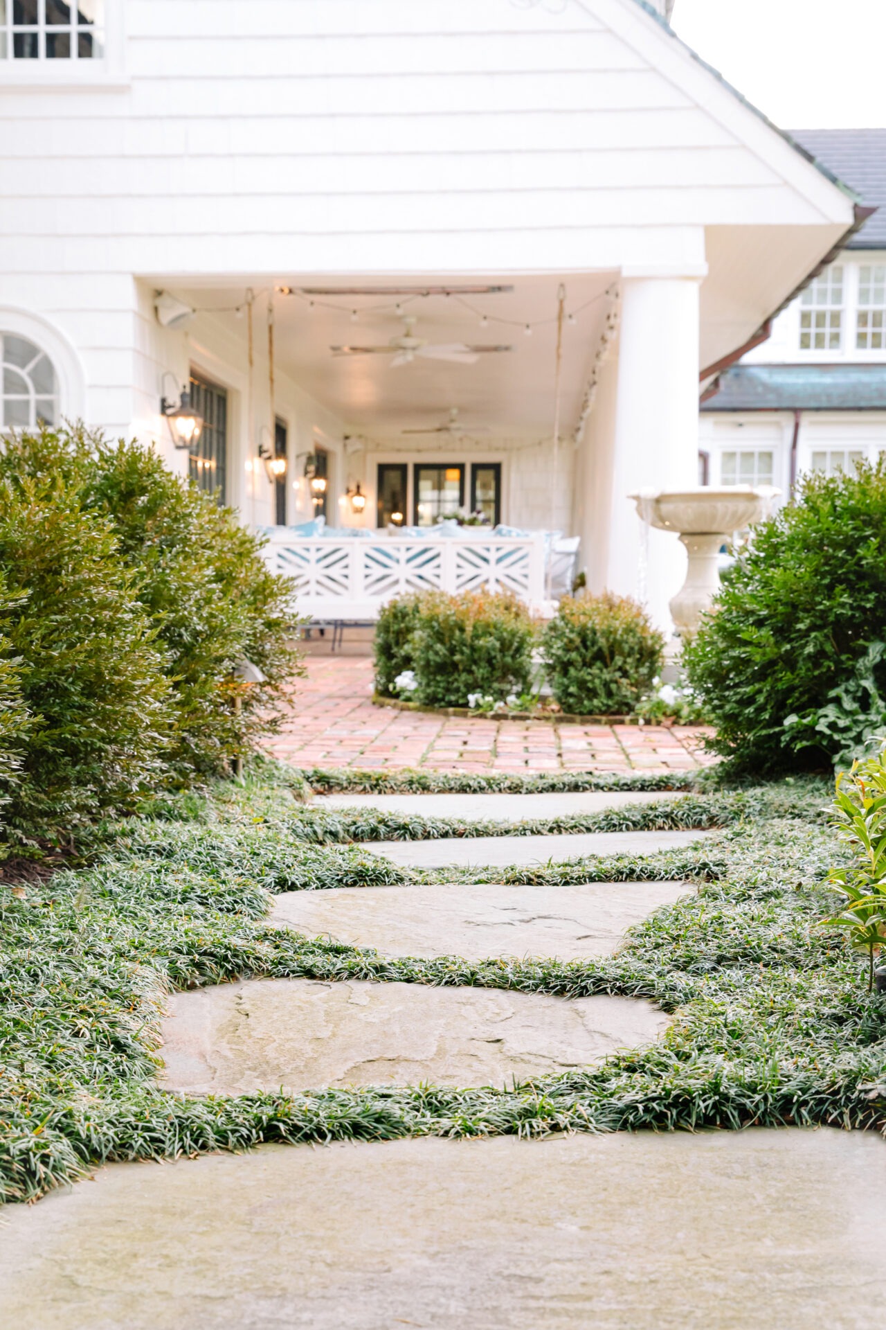 Stone pathway leads to a cozy porch with white railing, surrounded by manicured shrubs and warm lighting under an overhang of a house.