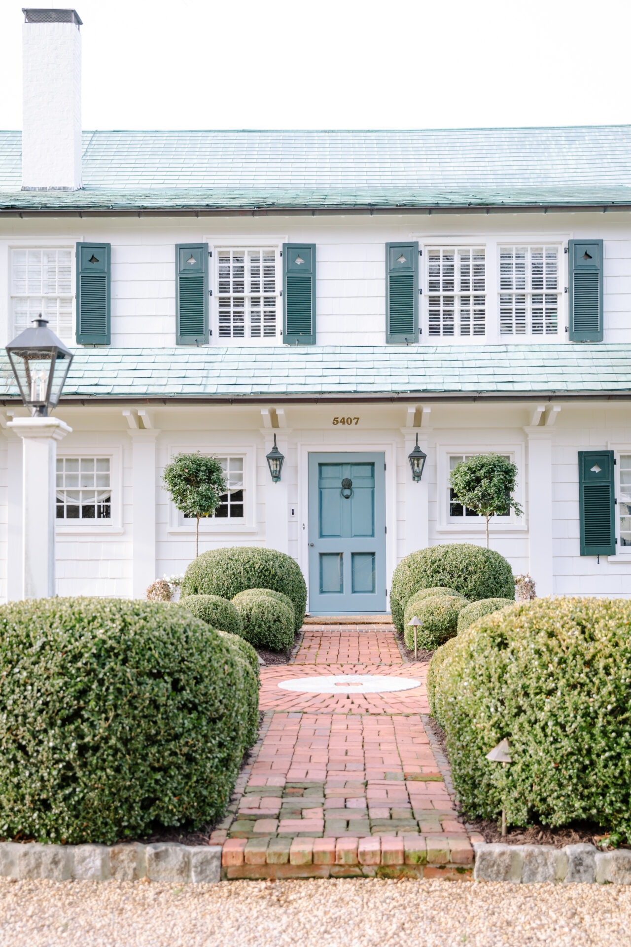 Charming house with a blue door, green shutters, and manicured bushes lining a brick walkway. Elegant, symmetrical facade evokes timeless elegance.