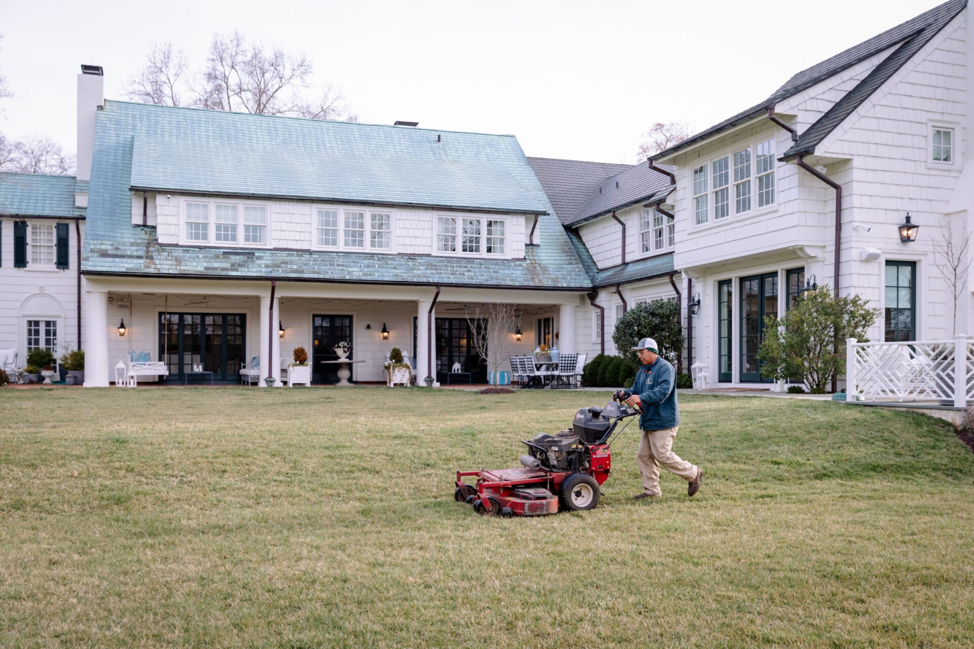 A person mowing the lawn in front of a large, traditional-style house with green roofing and white walls, surrounded by a spacious yard.