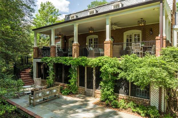 Two-story brick house with a large porch, surrounded by lush greenery. Outdoor seating on a stone patio enhances its charming, serene ambiance.