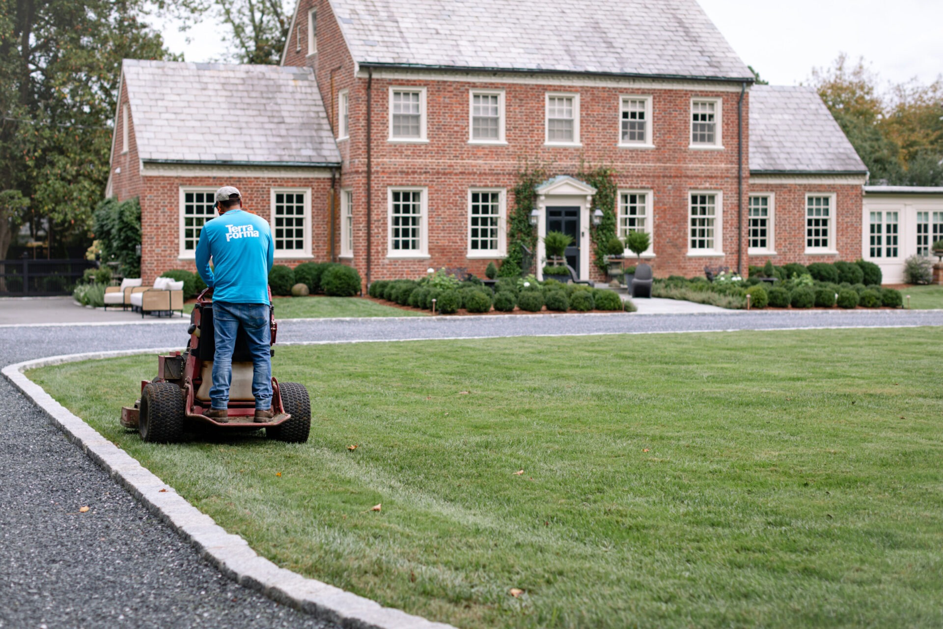 A person operates a lawnmower on a neatly trimmed lawn in front of a large, classic red brick house with white-framed windows.