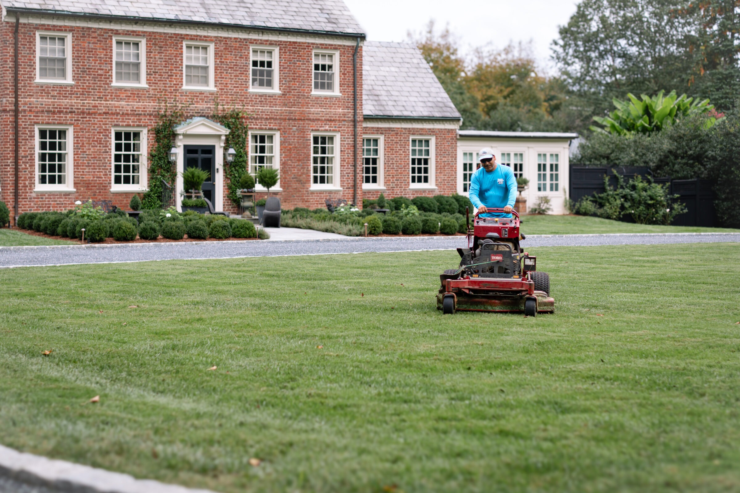 A person operates a lawnmower on a large green lawn in front of a brick house with multiple windows and a manicured garden.