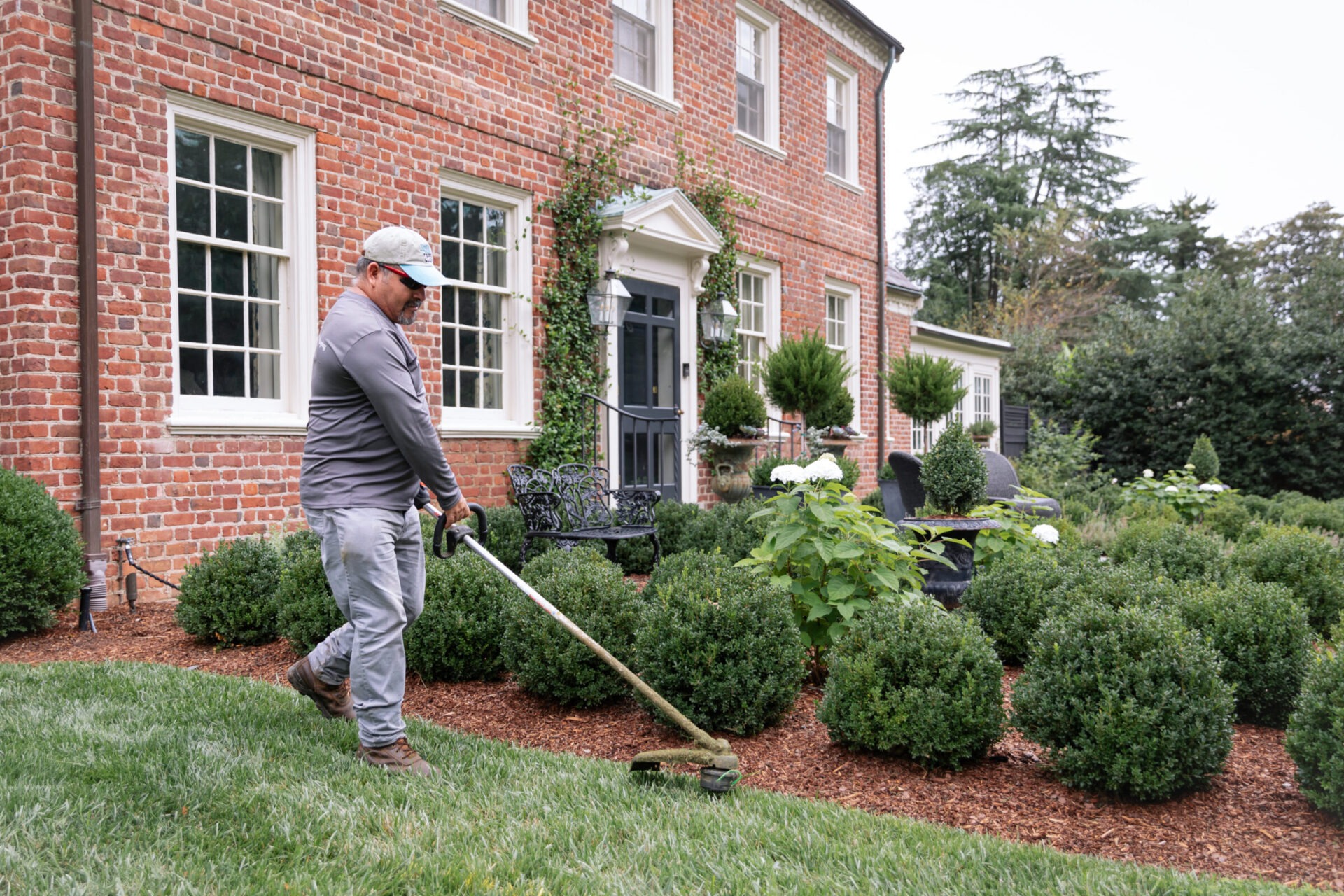 A person trims grass in front of a brick house with neatly maintained hedges and a decorative entrance, surrounded by greenery.