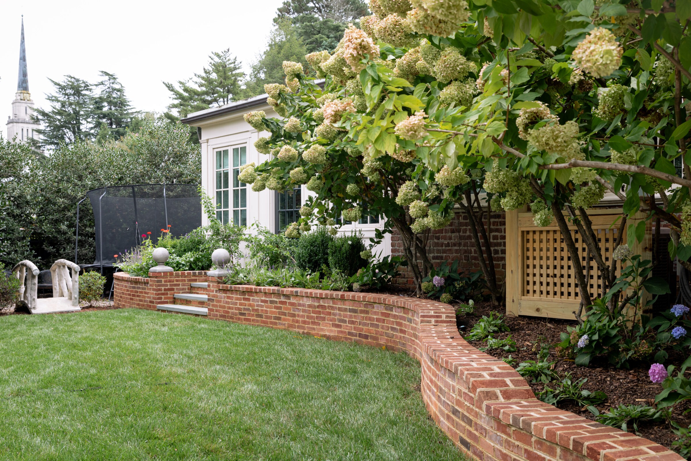 Lush garden with a curved brick wall, large hydrangeas, and a white house. A small bridge and a church spire are visible.