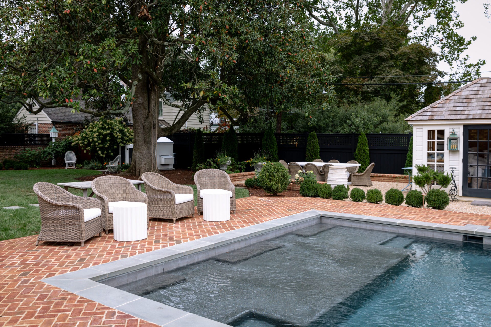 A spacious backyard with a pool, wicker chairs, brick patio, surrounded by greenery and trees, adjacent to a small white garden shed.