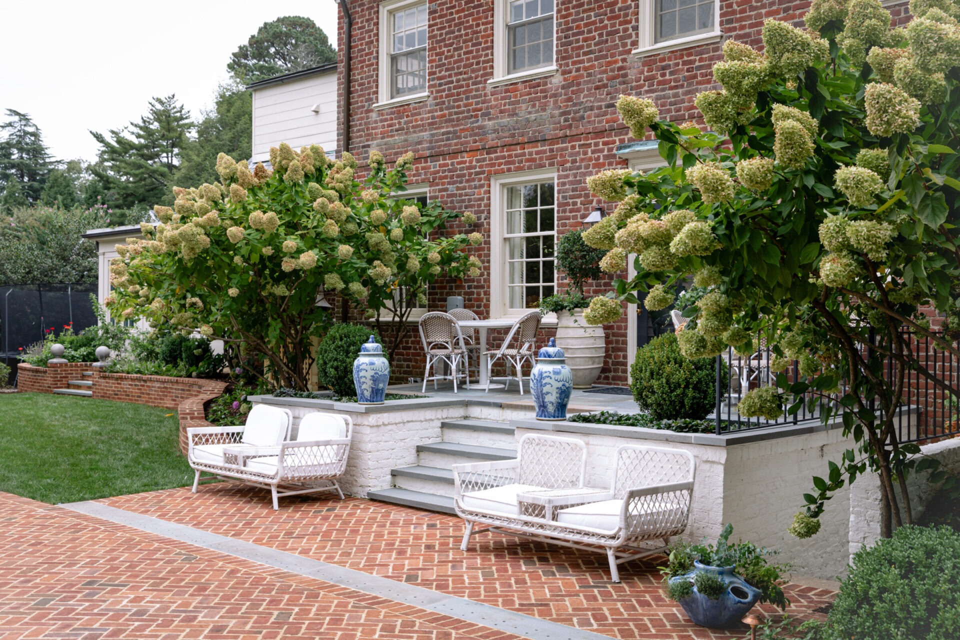 A charming garden patio features white seating, vibrant greenery, and hydrangea-lined steps leading to a brick house with elegant outdoor decor.