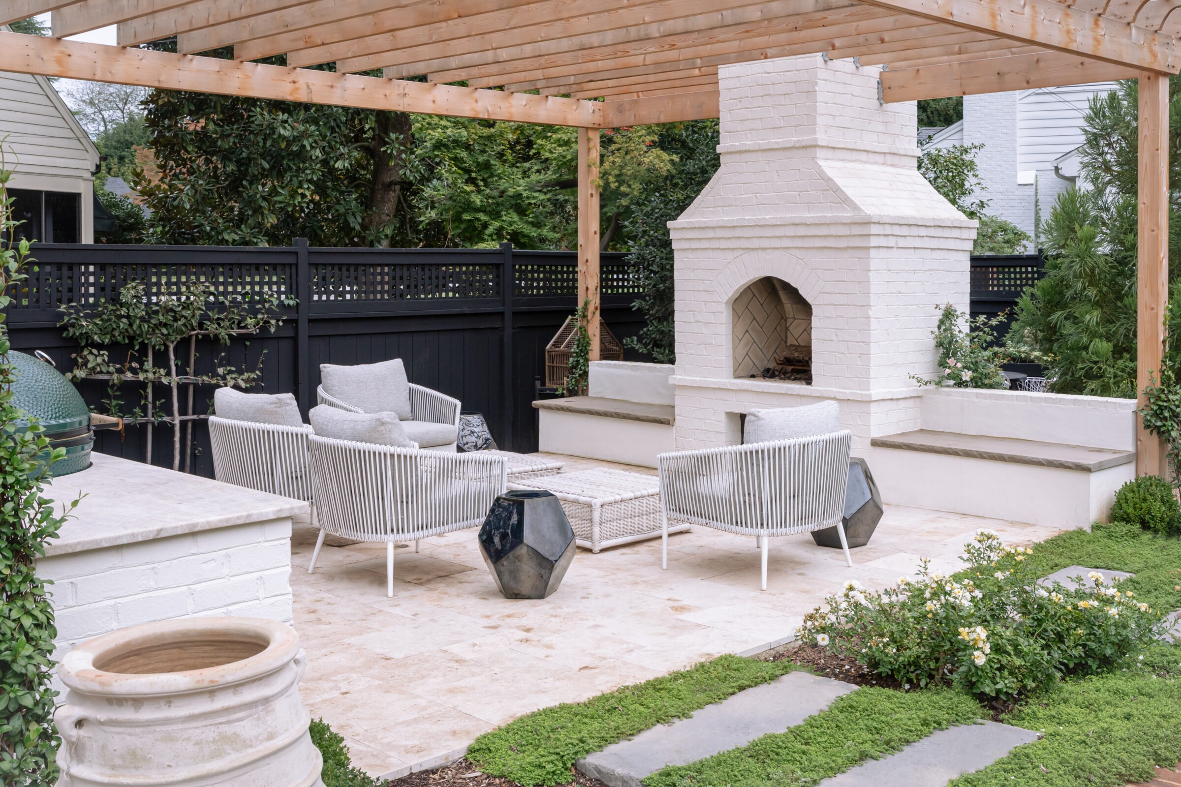 A modern outdoor patio with a white brick fireplace, pergola, grey seating, geometric tables, and landscaped garden. No people or recognizable landmarks present.
