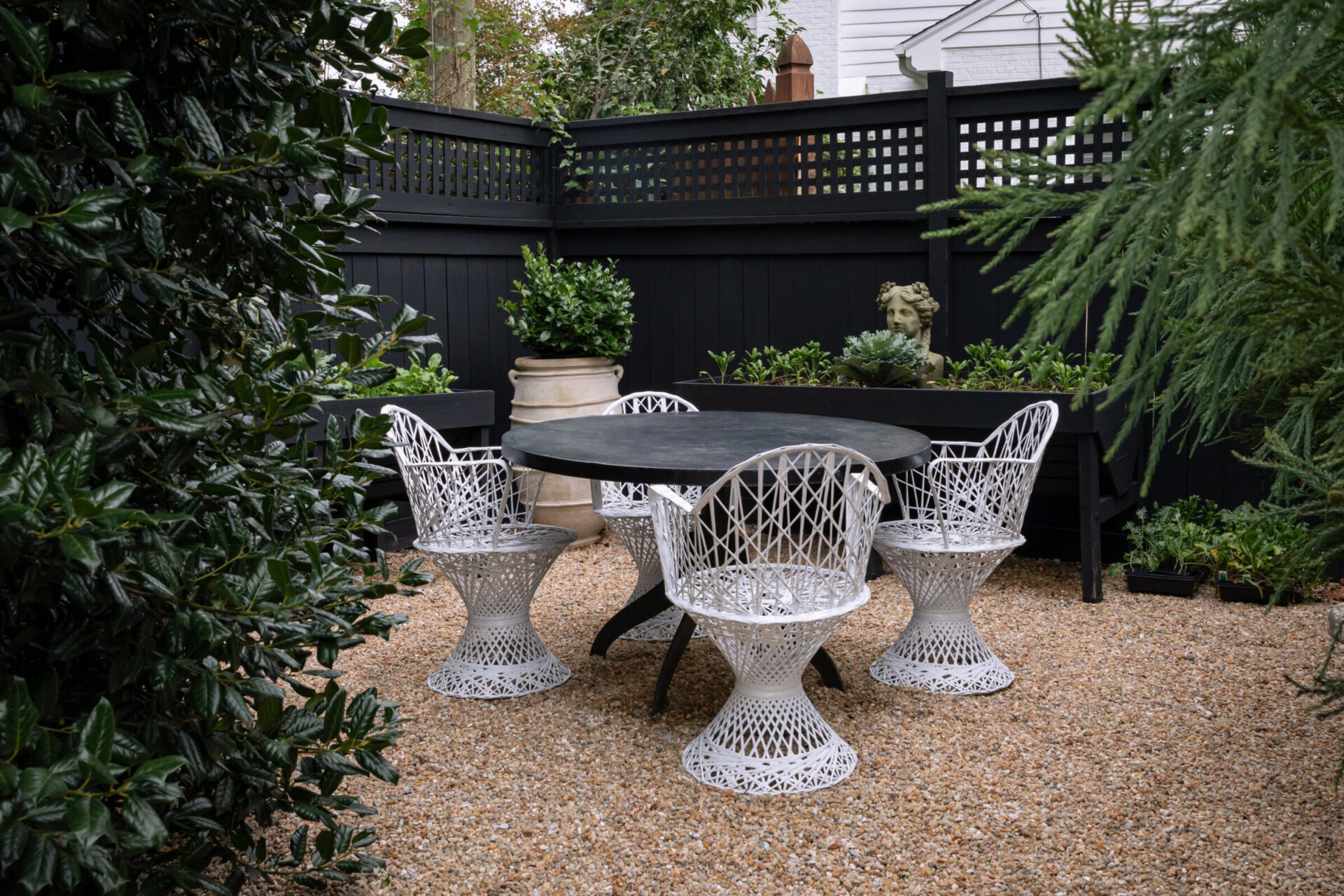 A cozy patio with white wicker chairs, a dark table, surrounded by greenery and tall black fencing in a private garden setting.