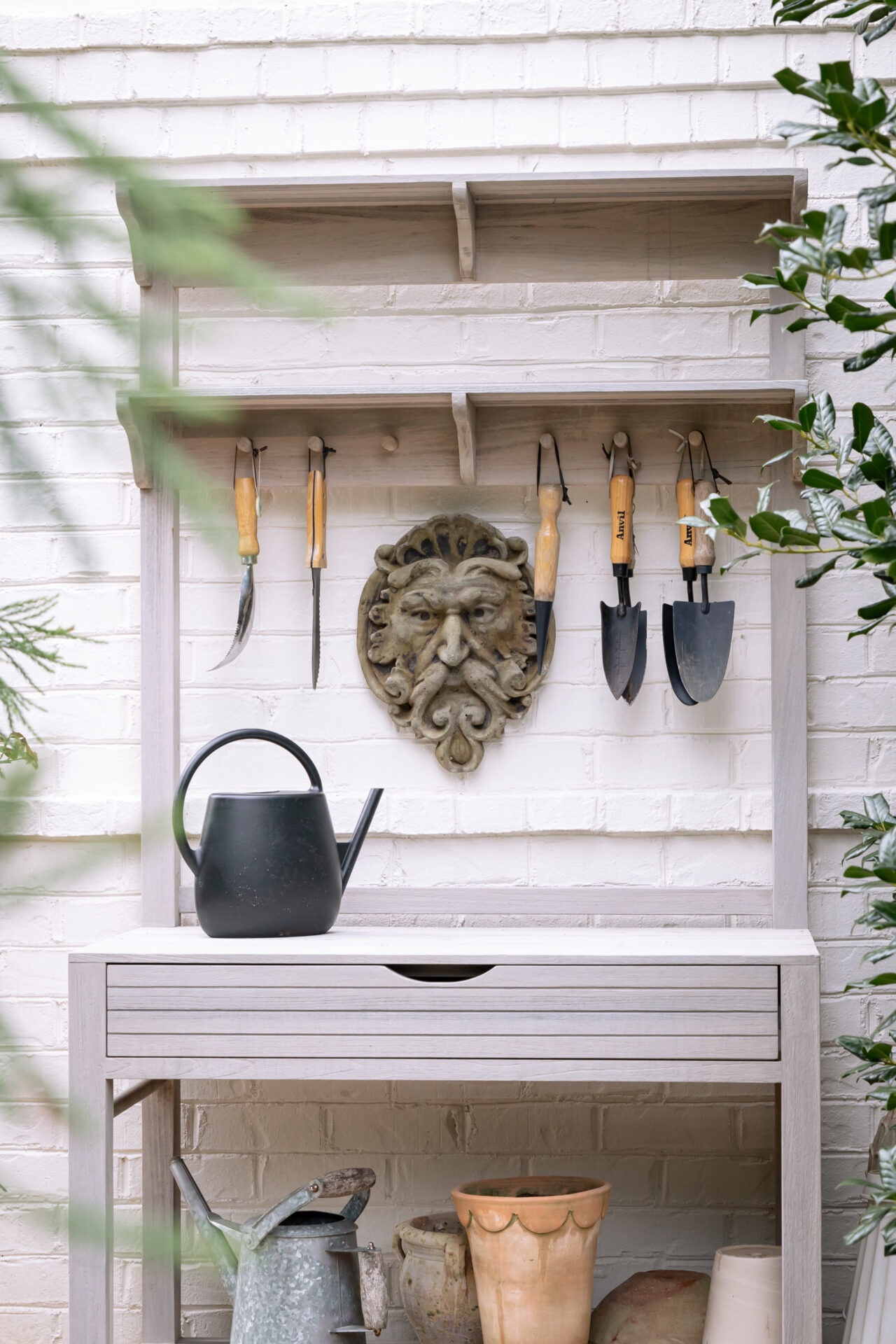 A garden workstation with hanging tools, classical face sculpture, black watering can, and flower pots against a white brick wall adorned with greenery.